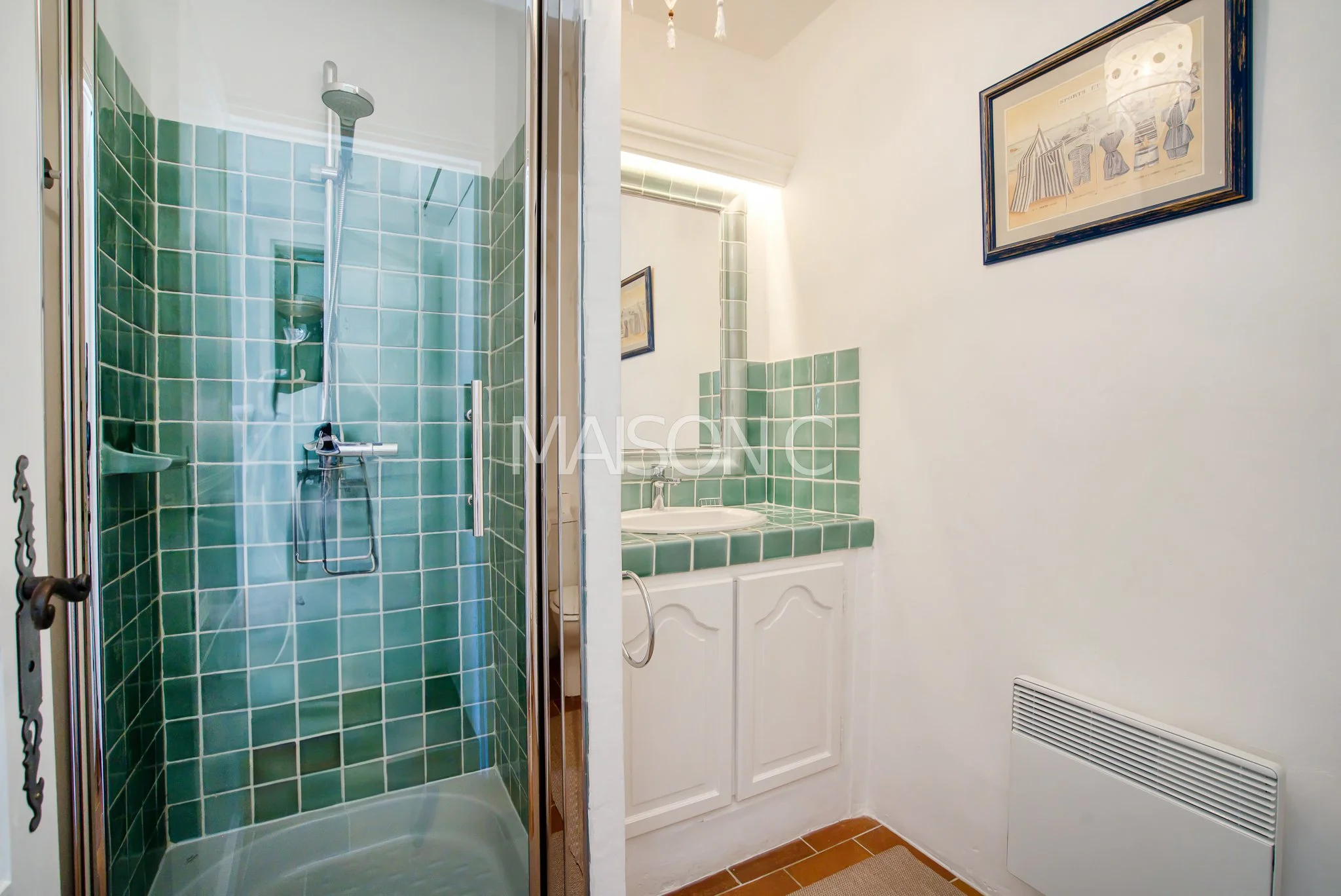 Salle de bain avec douche à l'italienne en carreaux verts, petit lavabo avec miroir et peinture encadrée sur le mur blanc.