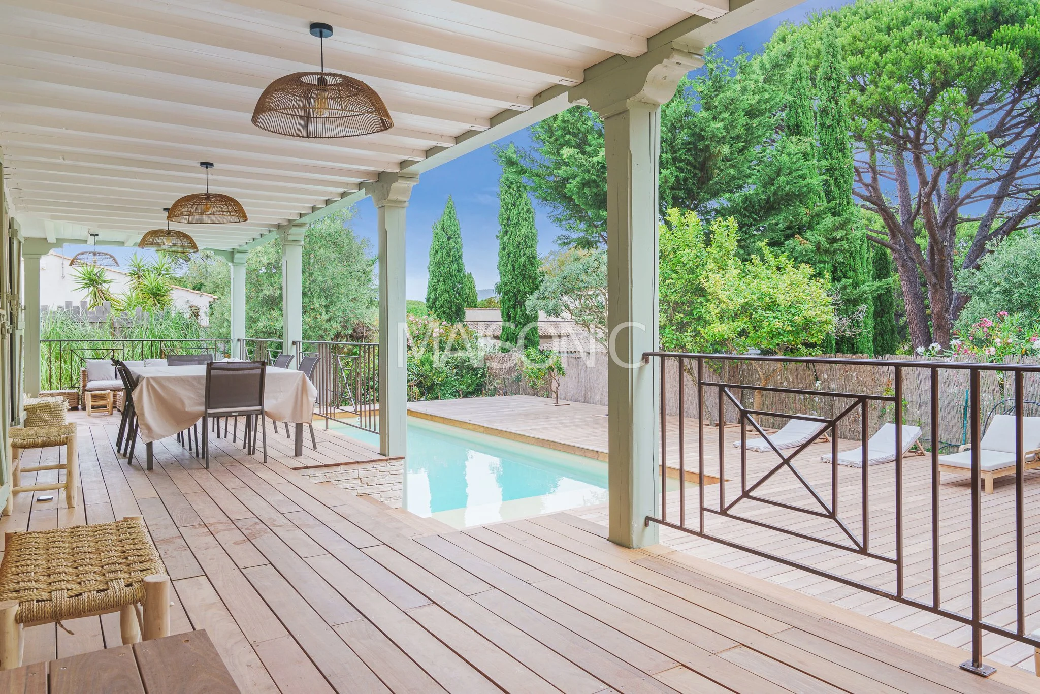 Terrasse en bois avec une table à manger, chaises, hamacs, piscine et jardin avec arbres et arbustes verts.