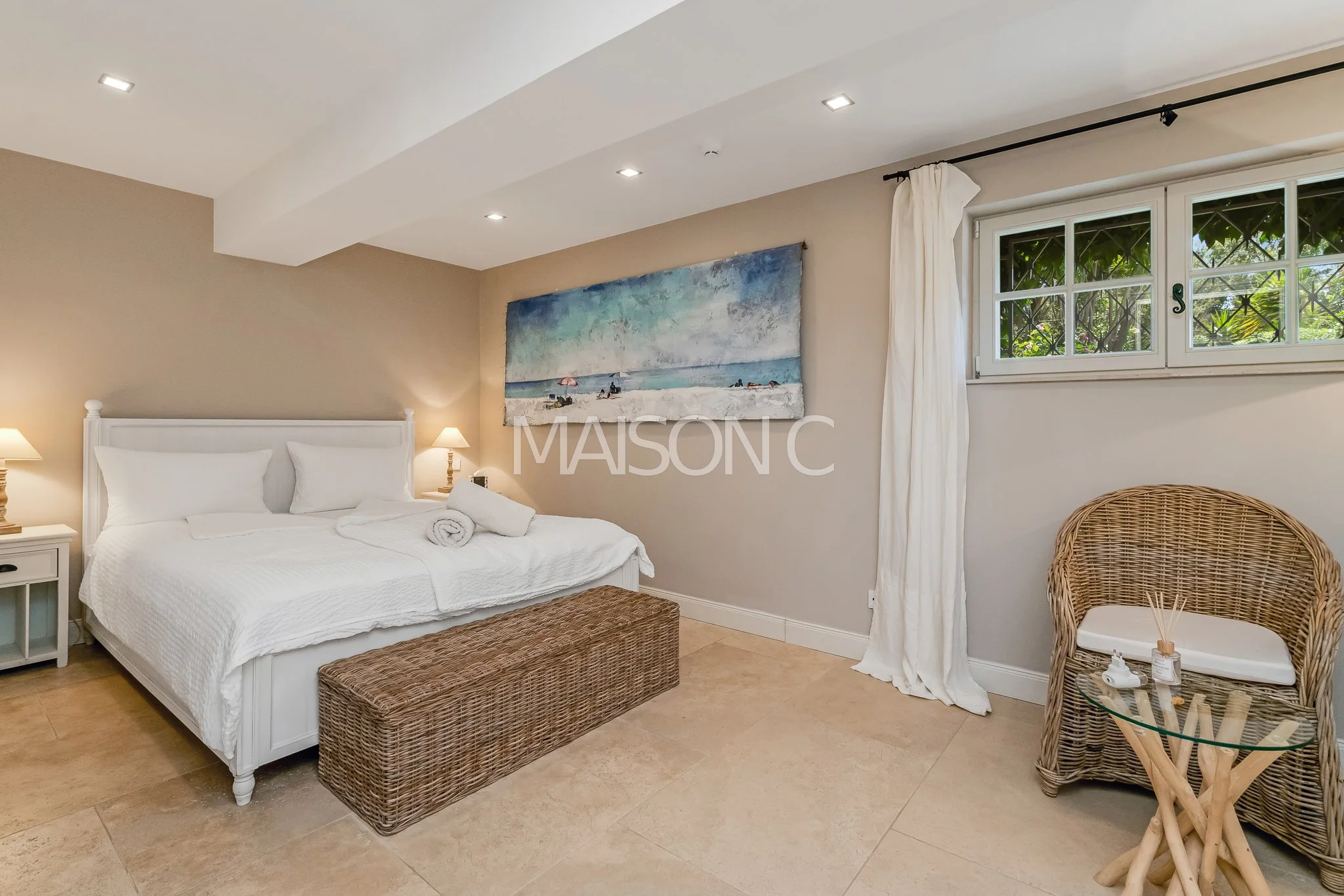 Chambre avec lit blanc, deux lampes de chevet, tableau de plage, fauteuil en rotin, fenêtre avec rideau blanc.