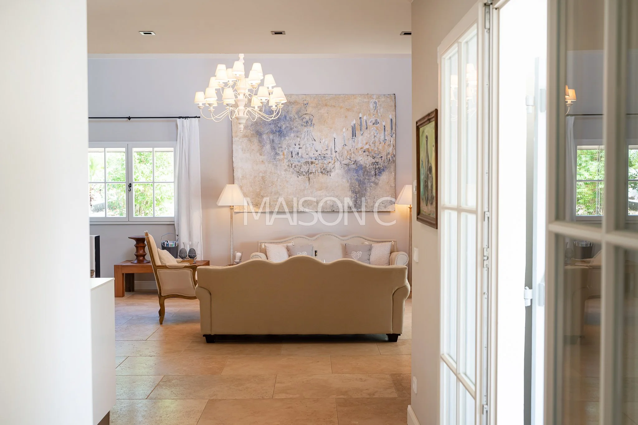 Salon lumineux avec canapé beige, tableau abstrait sur le mur, chandelier blanc, lampes de lecture, fenêtres ouvertes sur un jardin