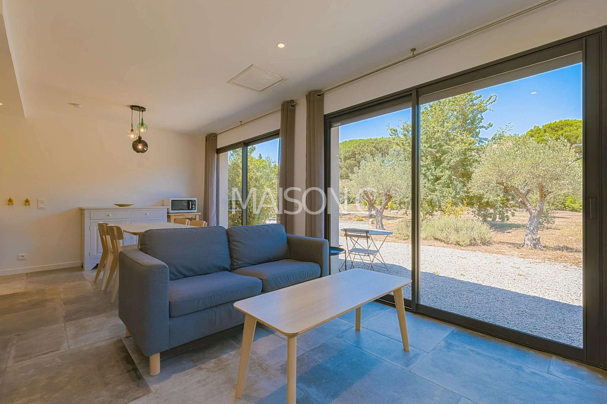Salon moderne avec canapé bleu, table en bois, grandes fenêtres donnant sur un jardin avec des arbres et un ciel bleu.