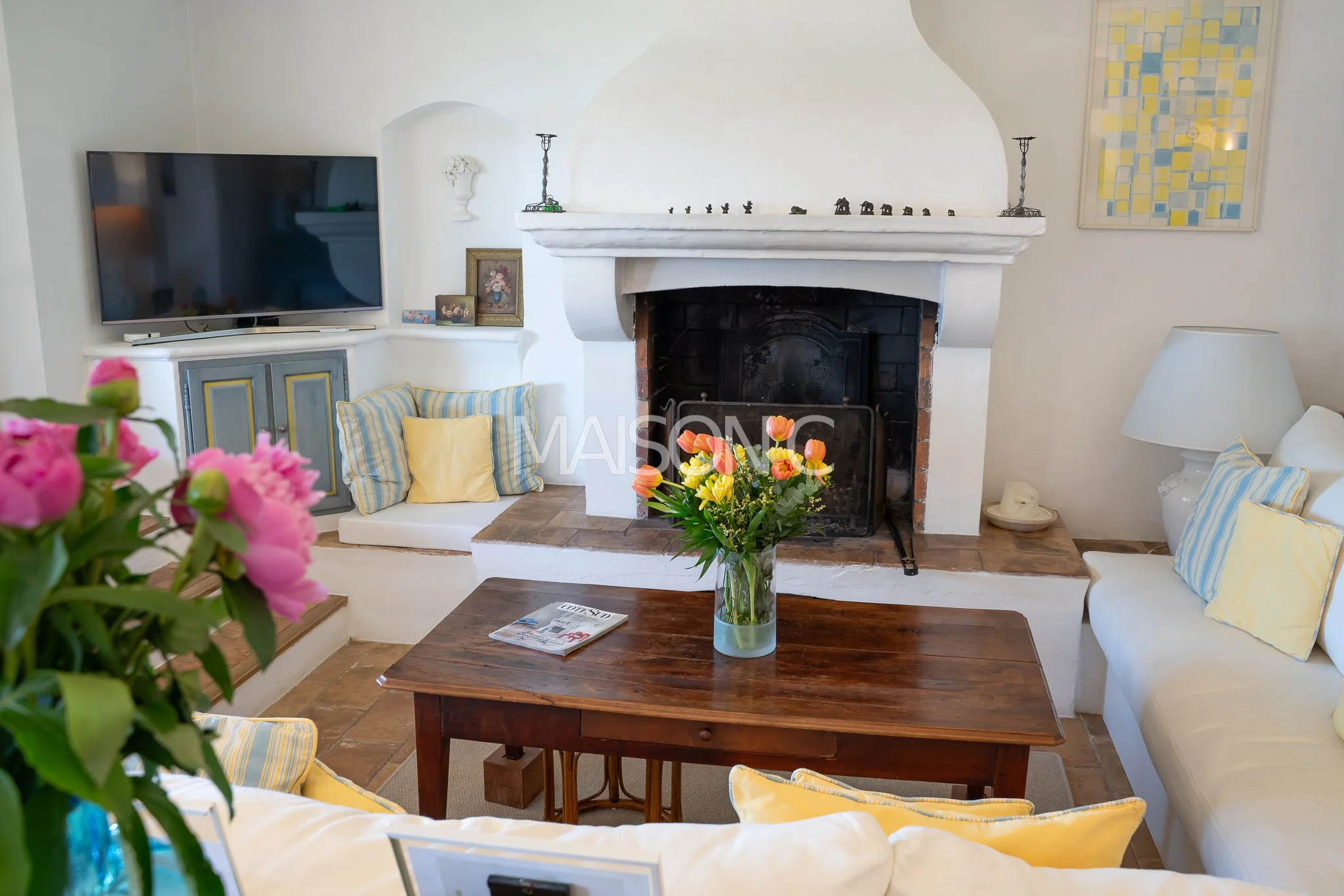 Salon avec cheminée en pierre, fleurs dans un vase sur une table en bois, télévision, canapé blanc avec coussins jaunes et bleus, peinture abstraite, lampadaire et décorations sur la cheminée.