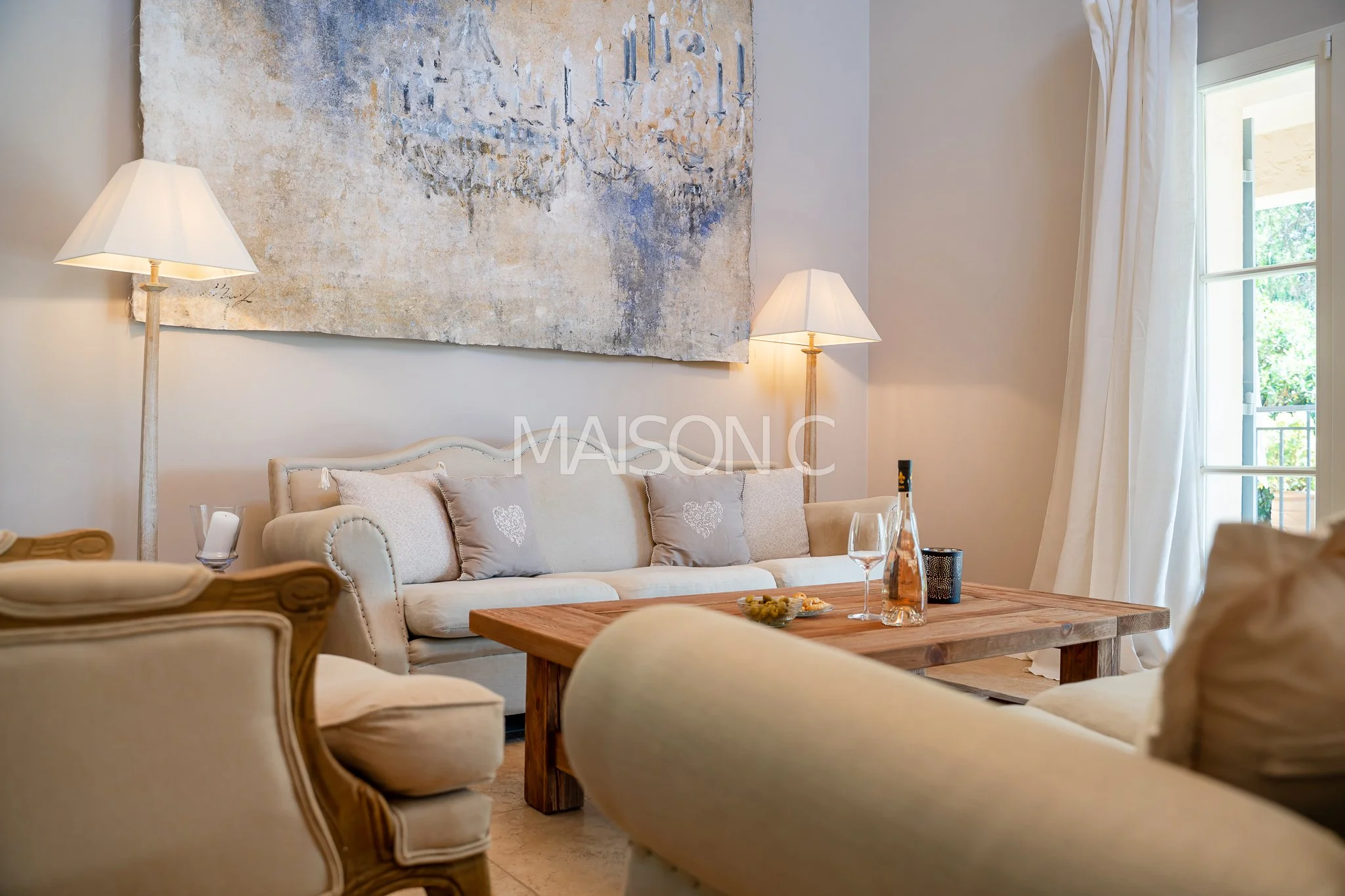 Salon avec canapé beige, tapisseries, lampes sur pied, table en bois, verre de vin, bouteille d'eau, et portes-fenêtres avec rideaux blancs.