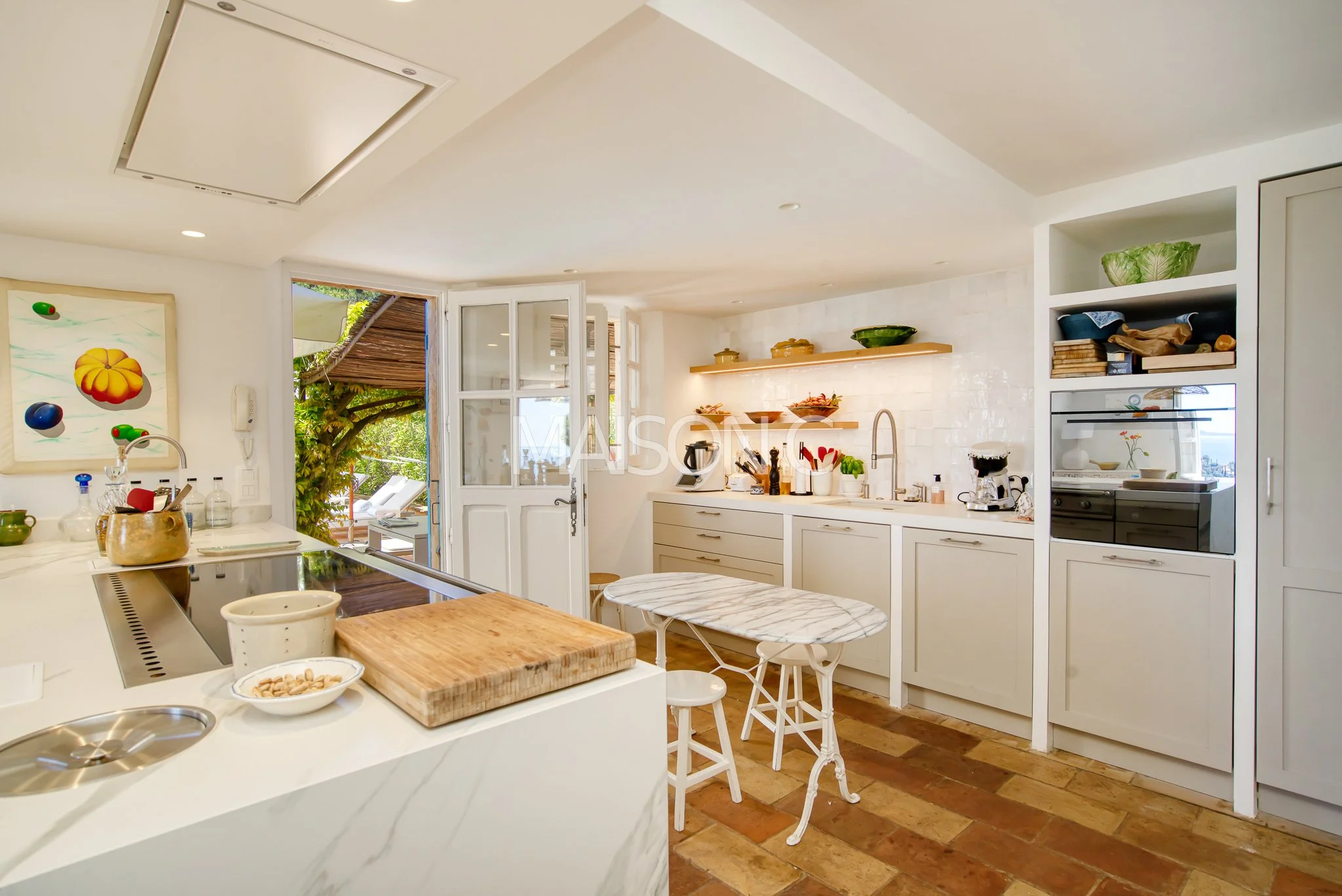 Cuisine lumineuse avec comptoir en marbre, plaques de cuisson, étagères avec vaisselle et pots de cuisine, porte-fenêtre ouvrant sur un espace extérieur avec des chaises longues et des parasols.