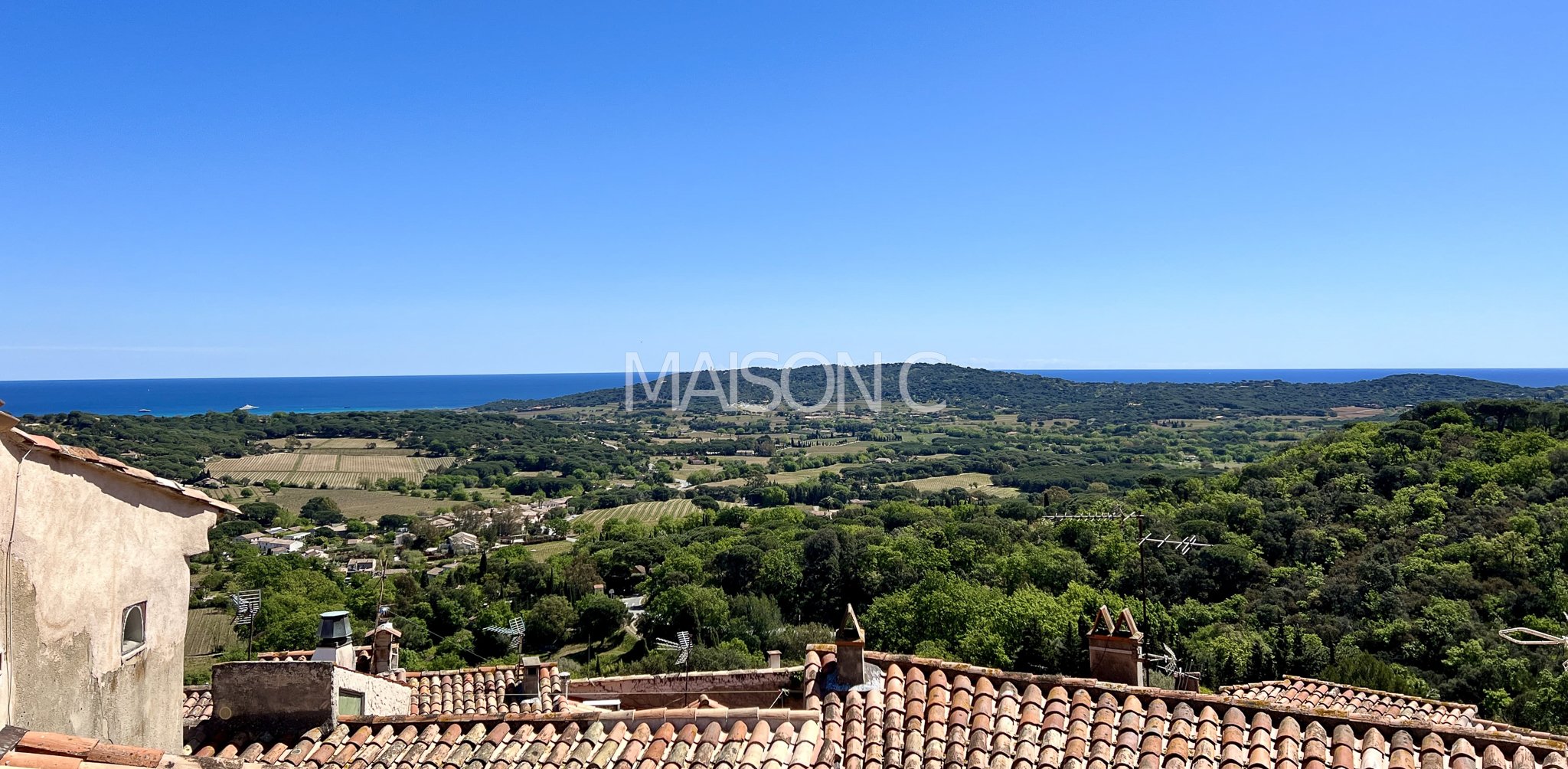 Paysage de campagne avec des tuiles en terre cuite au premier plan, des arbres verts, des champs cultivés, une colline et la mer au loin sous un ciel bleu clair.