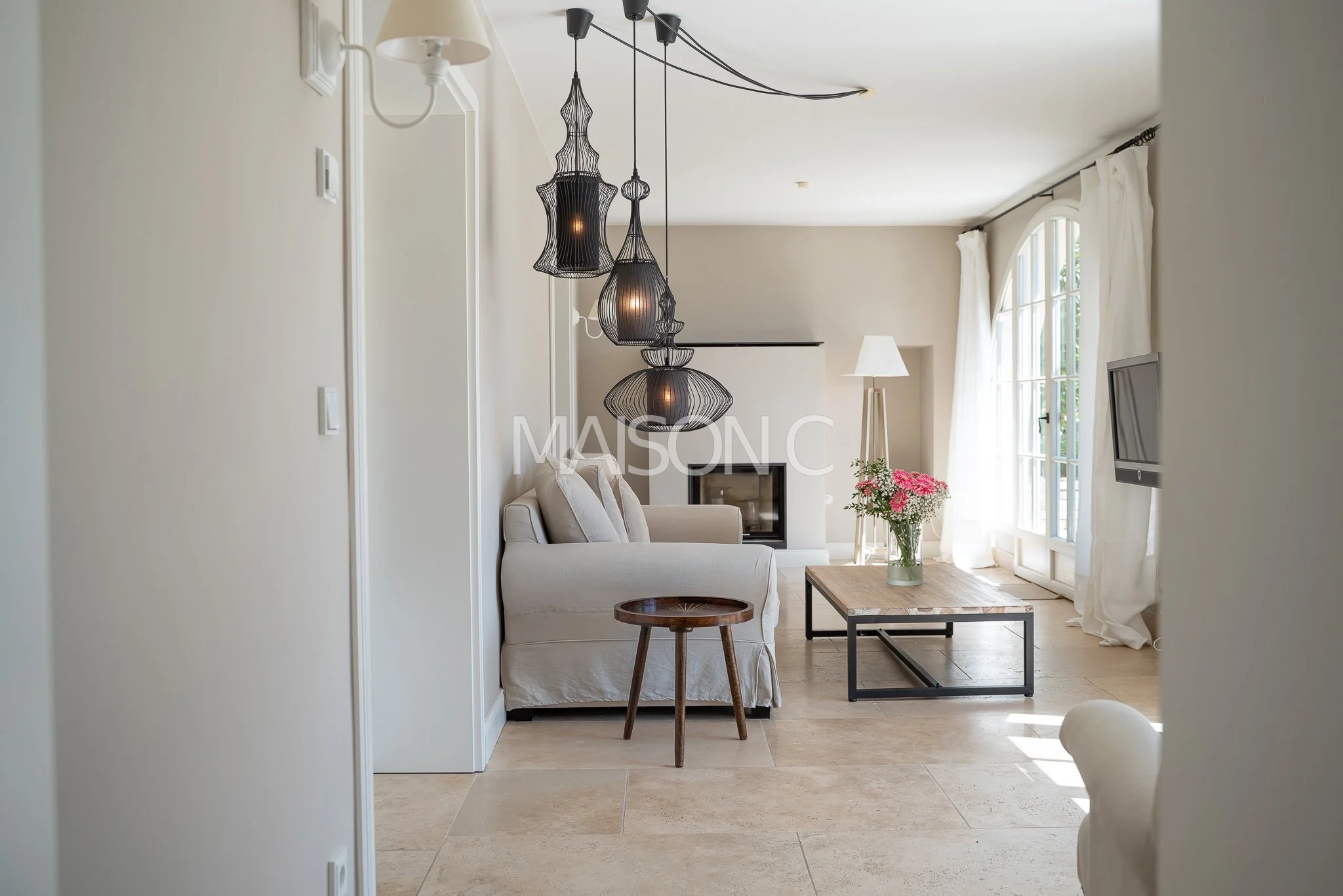 Salon contemporain avec canapé beige, table en bois, lampes suspendues noire, cheminée et télévision, grandes fenêtres avec rideaux blancs, fleurs roses dans un vase, lumière naturelle