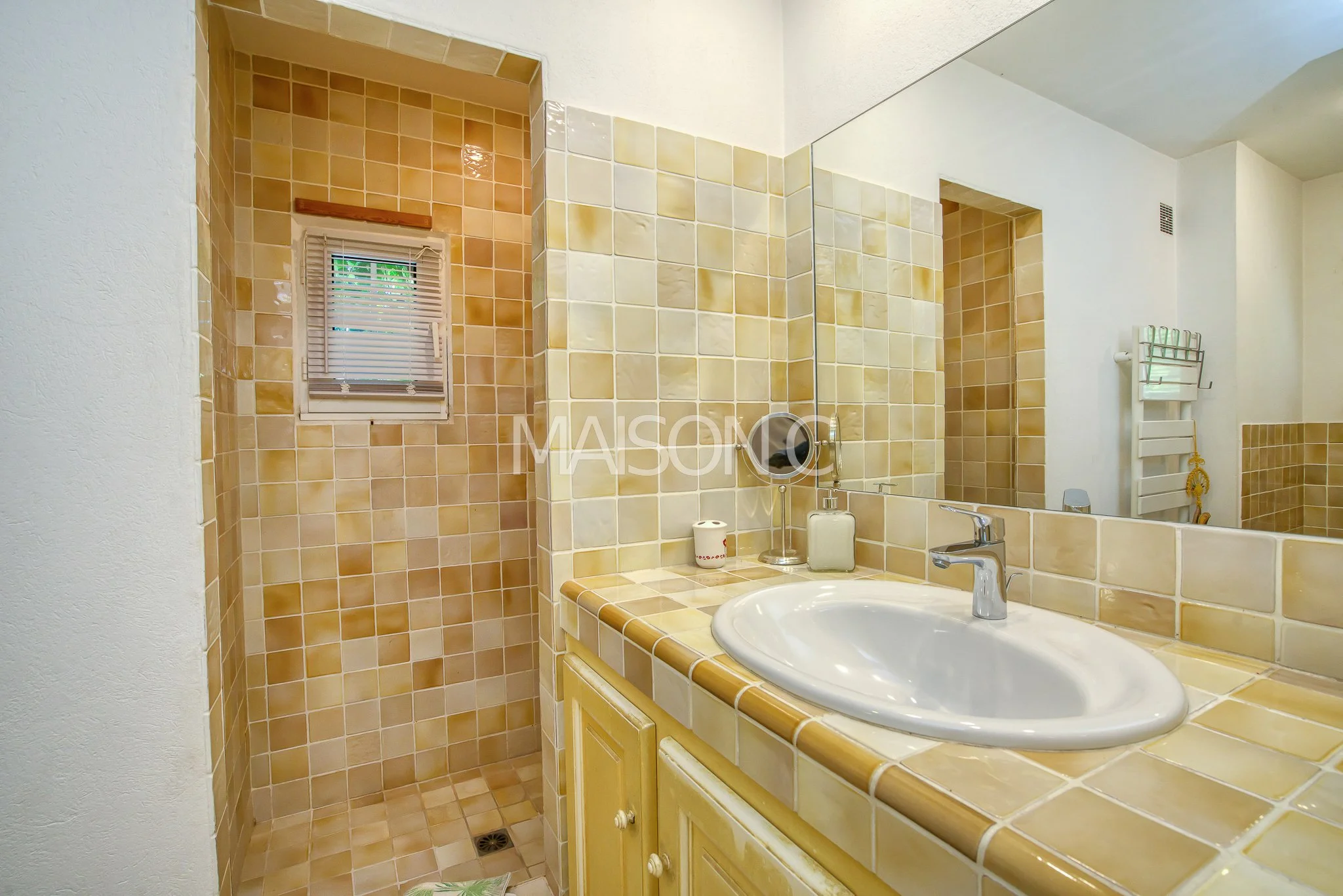 Salle de bain avec un lavabo en céramique blanc, un grand miroir, des carreaux beiges au mur et au comptoir, et une douche avec un petit fenêtre à volet blanc.