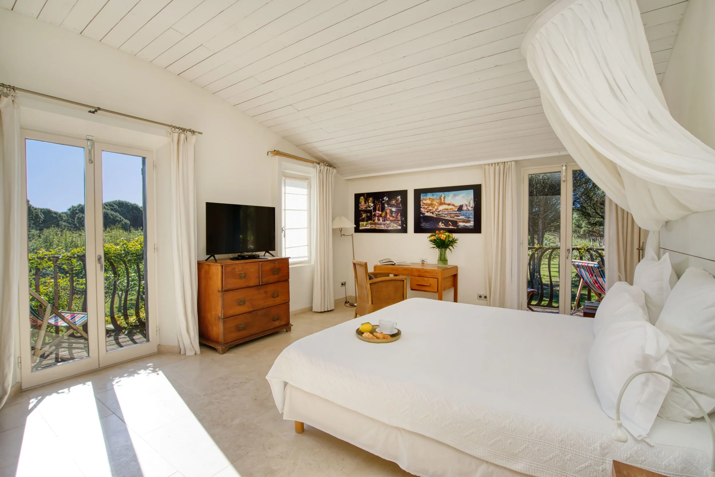Chambre lumineuse avec lit blanc, télévision, toilette, et balcon avec vue sur la nature.