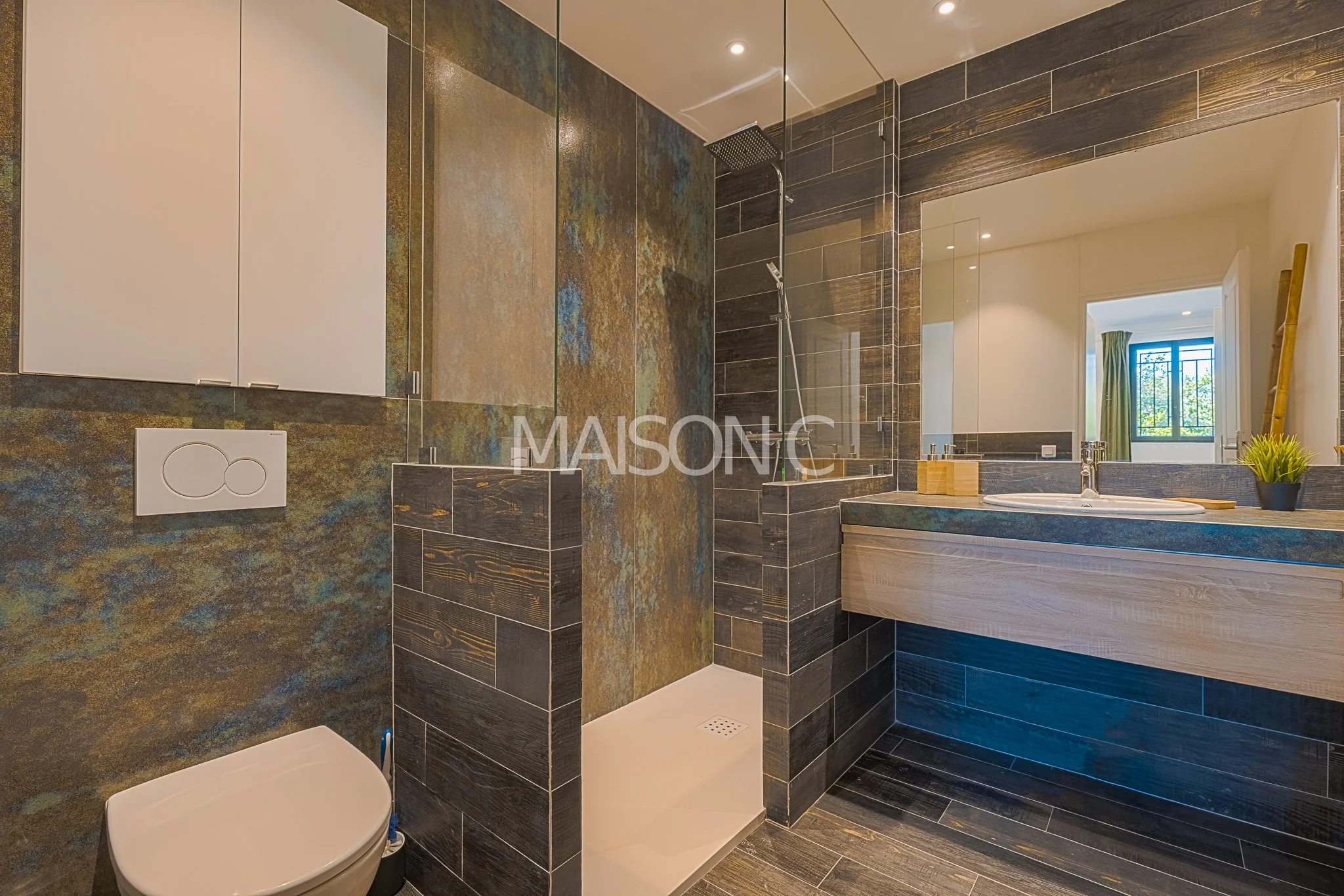 Salle de bain moderne avec un espace de douche, un WC suspendu, un lavabo avec miroir et un grand miroir mural, des murs et un sol recouverts de carreaux noirs et marron, fenêtre laissant entrer la lumière naturelle.