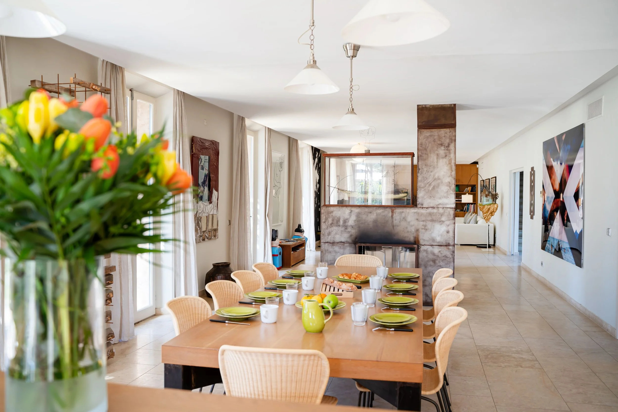 Salle à manger avec une grande table, assiettes vertes, tasses blanches, verres, fruits et repas, dans une pièce lumineuse avec des fenêtres et des œuvres d'art.