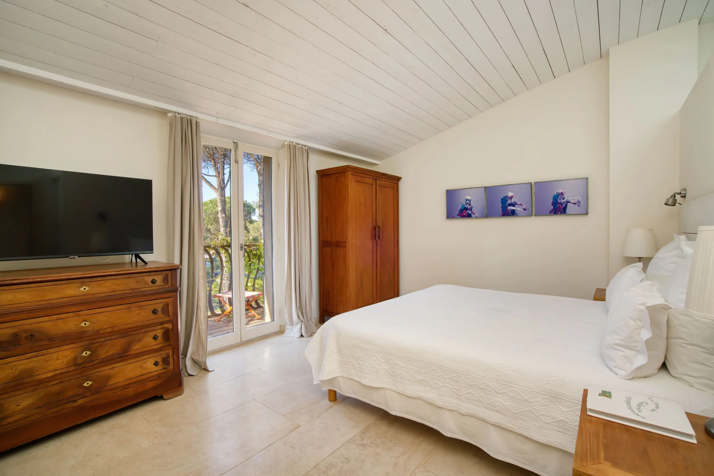 Chambre confortable avec un lit blanc, une armoire en bois, une commode en bois avec une télévision, une porte-fenêtre donnant sur un balcon, des rideaux beiges, une œuvre d'art moderne au-dessus du lit, et une lampe de chevet.