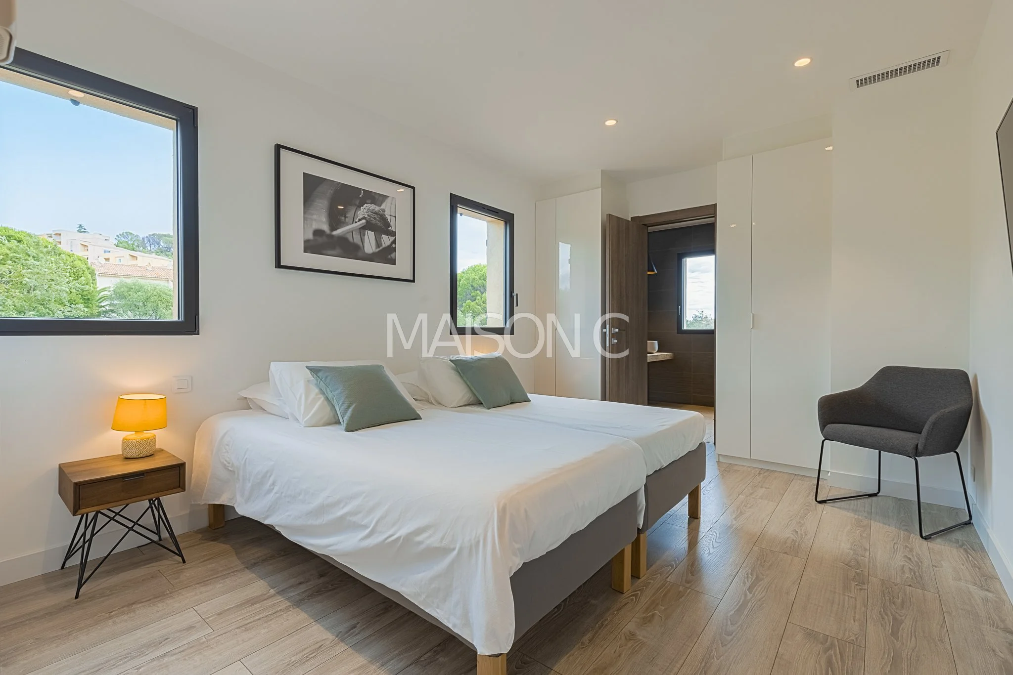 Chambre moderne avec lit double, deux fenêtres, une petite table de nuit avec lampe, un fauteuil gris, image en noir et blanc au mur, porte donnant sur salle de bain, décoration minimaliste