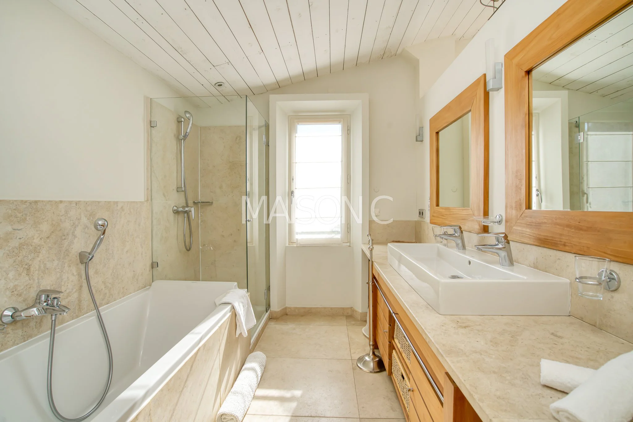 Salle de bain avec baignoire, douche à l'italienne, double lavabo avec miroirs en bois, fenêtre au centre, murs beige et plafond en bois clair.