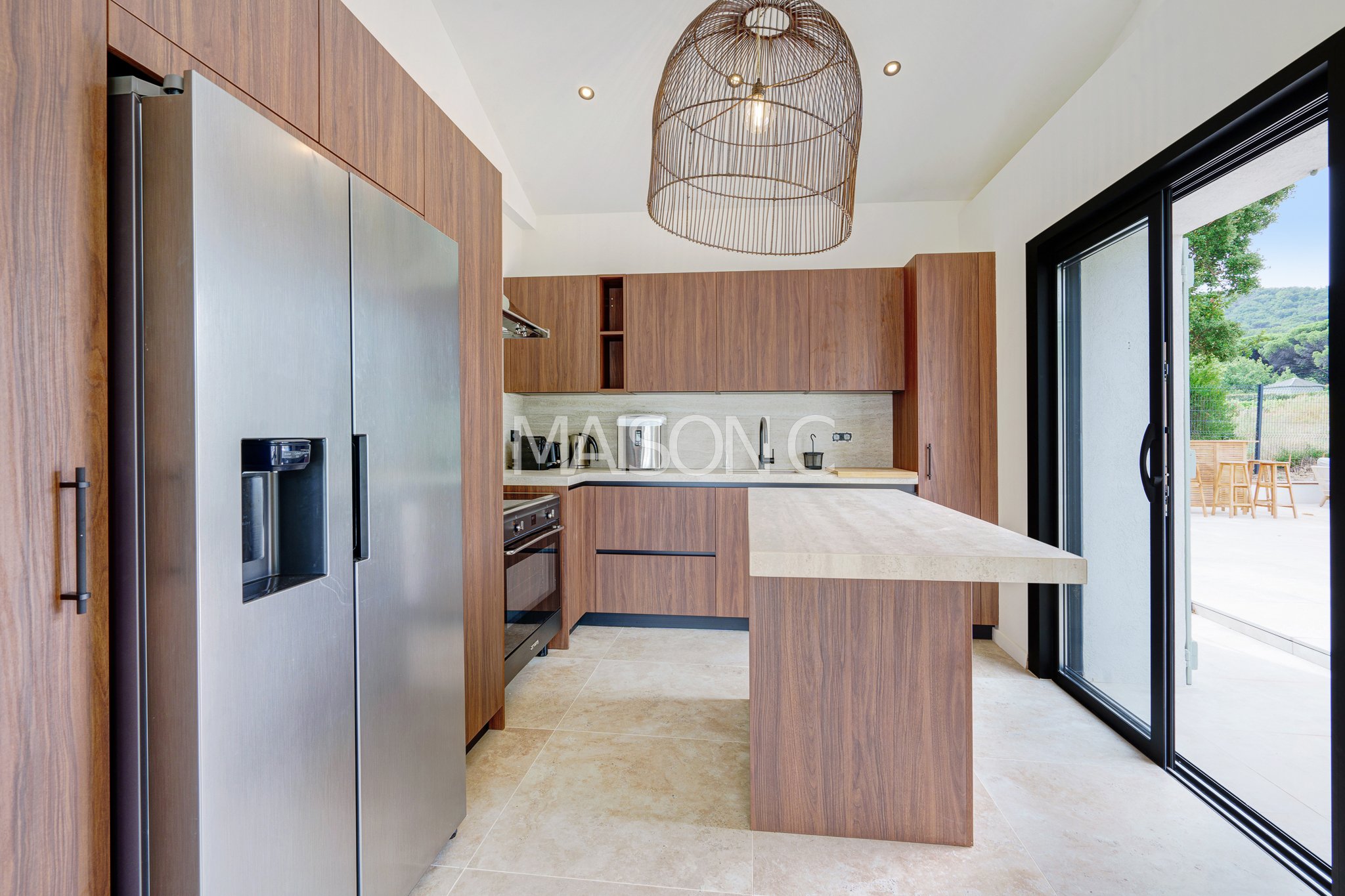 Cuisine moderne avec armoires en bois, réfrigérateur en acier inoxydable, carrelage beige, et porte-fenêtre donnant sur une terrasse extérieure.