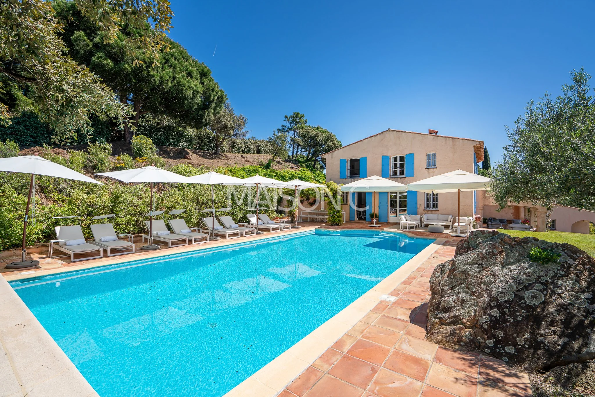 Piscine extérieure à proximité d'une maison beige avec volets bleus, entourée de chaises longues avec parasols blancs, dans un environnement de jardin avec arbres et rochers, par temps ensoleillé.