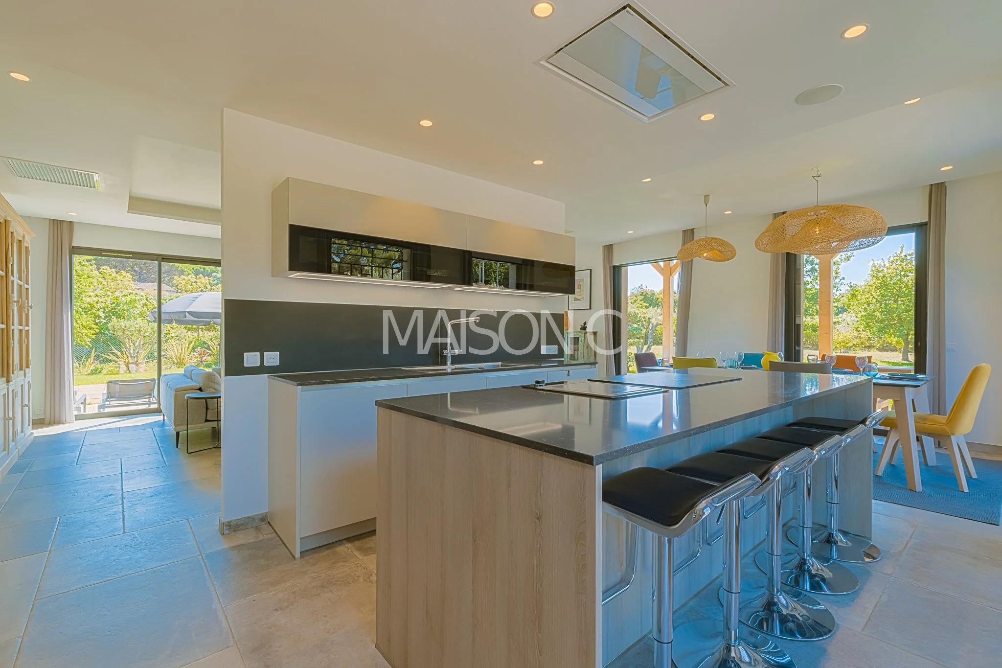 Cuisine moderne avec îlot central en bois, plan de travail noir, éclairage naturel par de grandes fenêtres, espace déjeuner avec chaises colorées, décoration épurée.