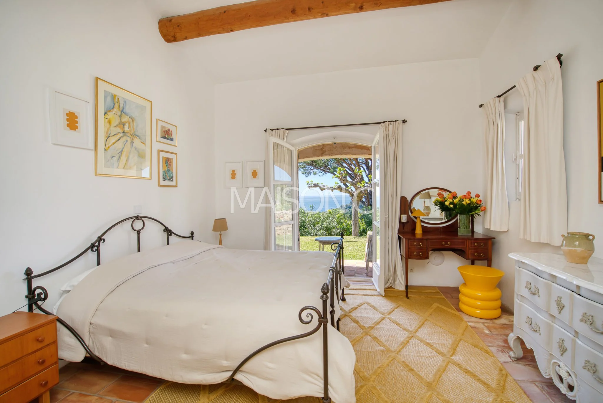 Chambre lumineuse avec un lit à cadre en métal, porte-fenêtre ouverte sur un jardin avec vue sur la mer, décoration colorée avec tableau, meubles en bois et pot de fleurs.