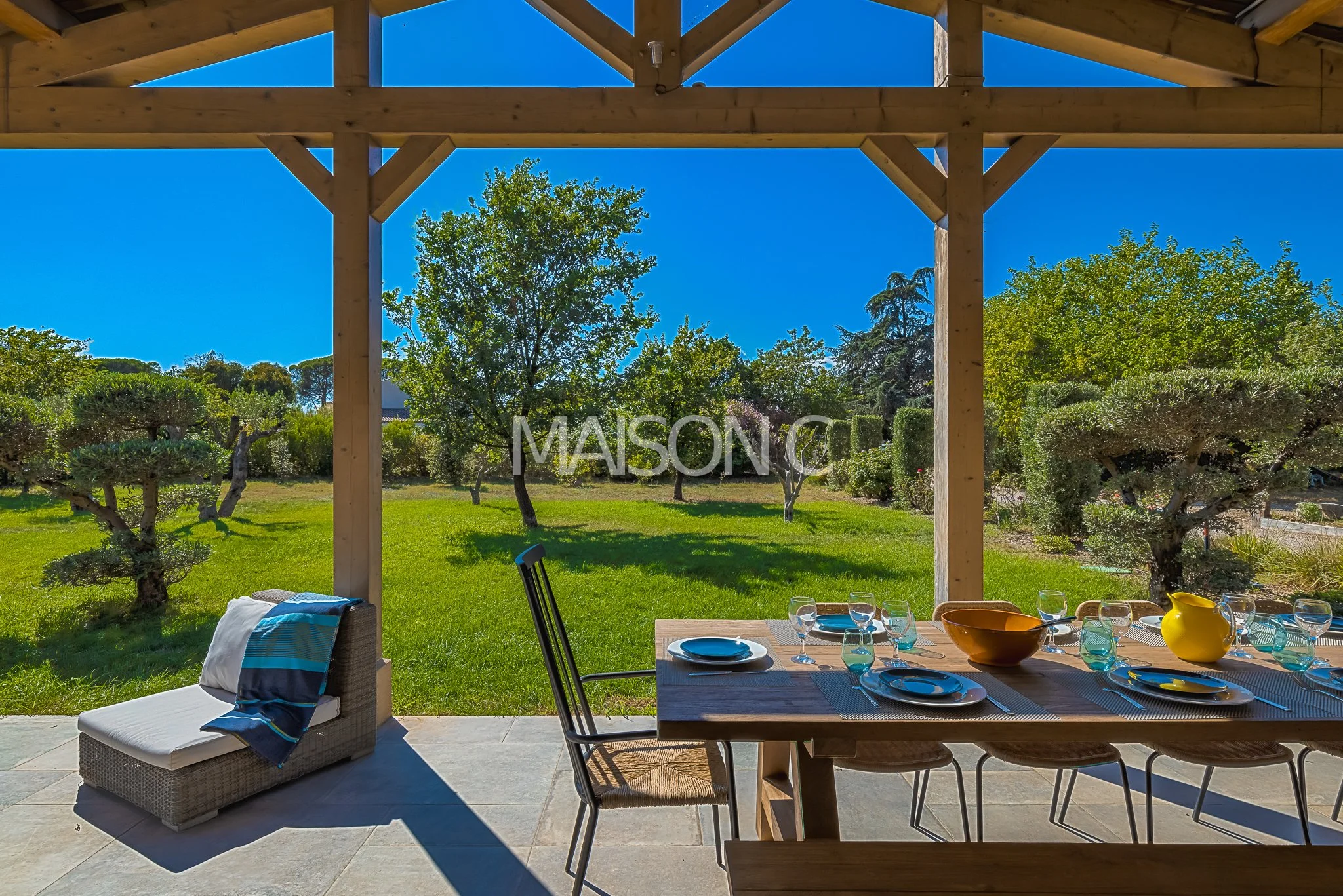 Table de nourriture avec assiettes, verres, carafe, bol et vase, vue sur un jardin avec des arbres et un ciel bleu
