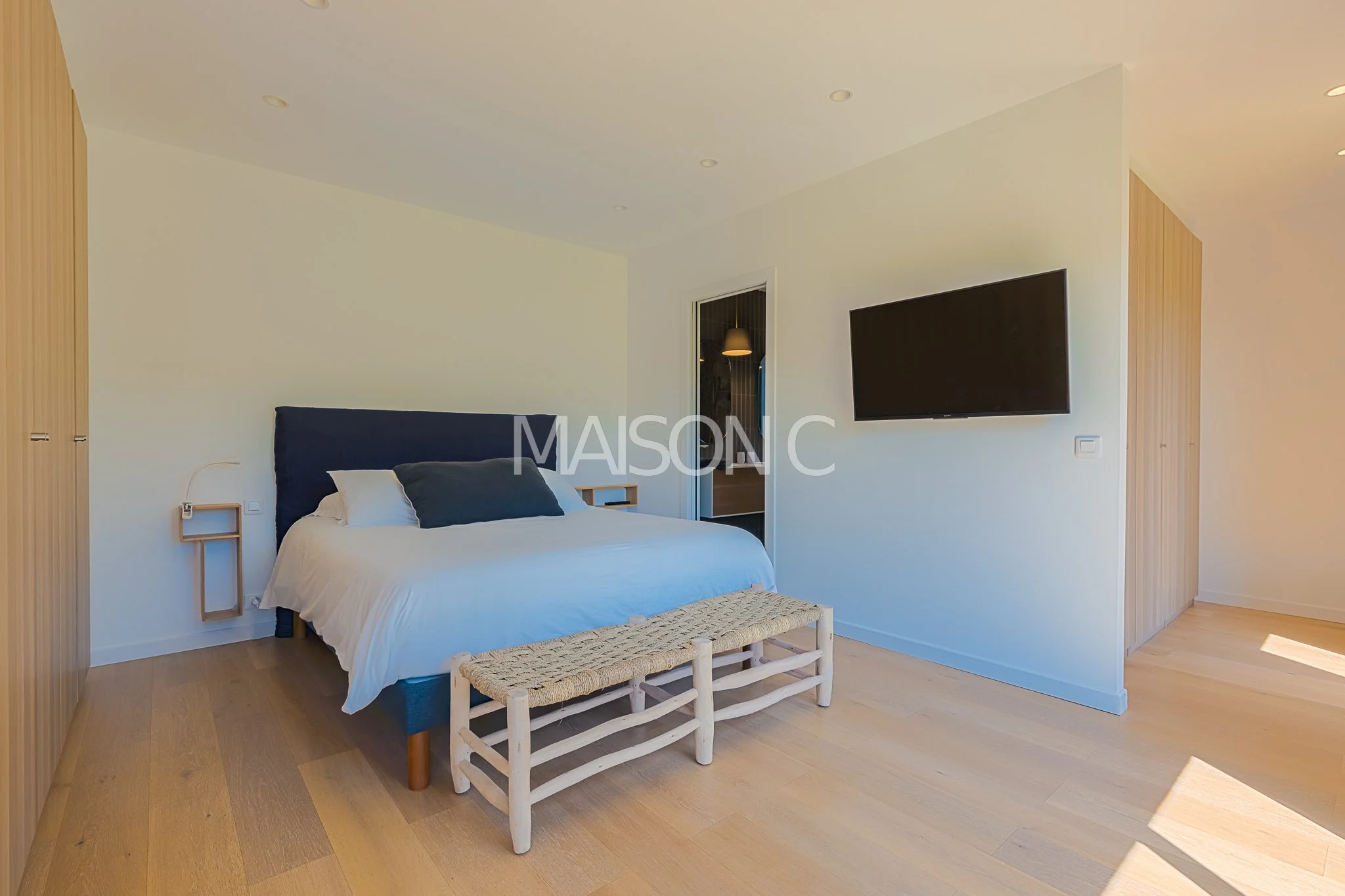 Chambre moderne avec lit, sol en bois clair, téléviseur mural, porte de salle de bain ouverte, meuble en bois naturel, mur blanc, ambiance épurée et lumineuse.
