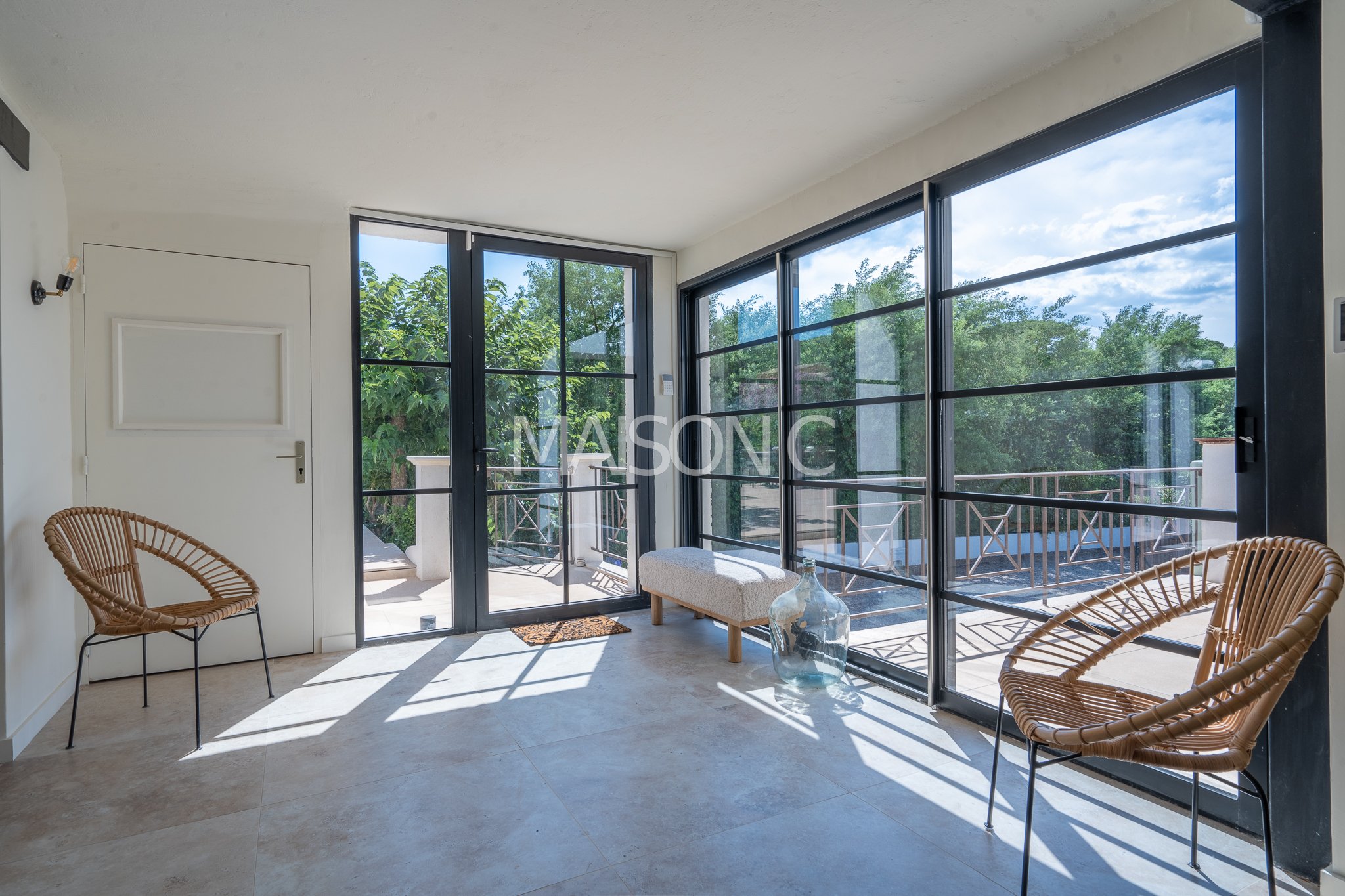 Salon lumineux avec grande baie vitrée, deux chaises en rotin, un banc rembourré, un grand vase en verre, vue sur un balcon et la verdure extérieure.