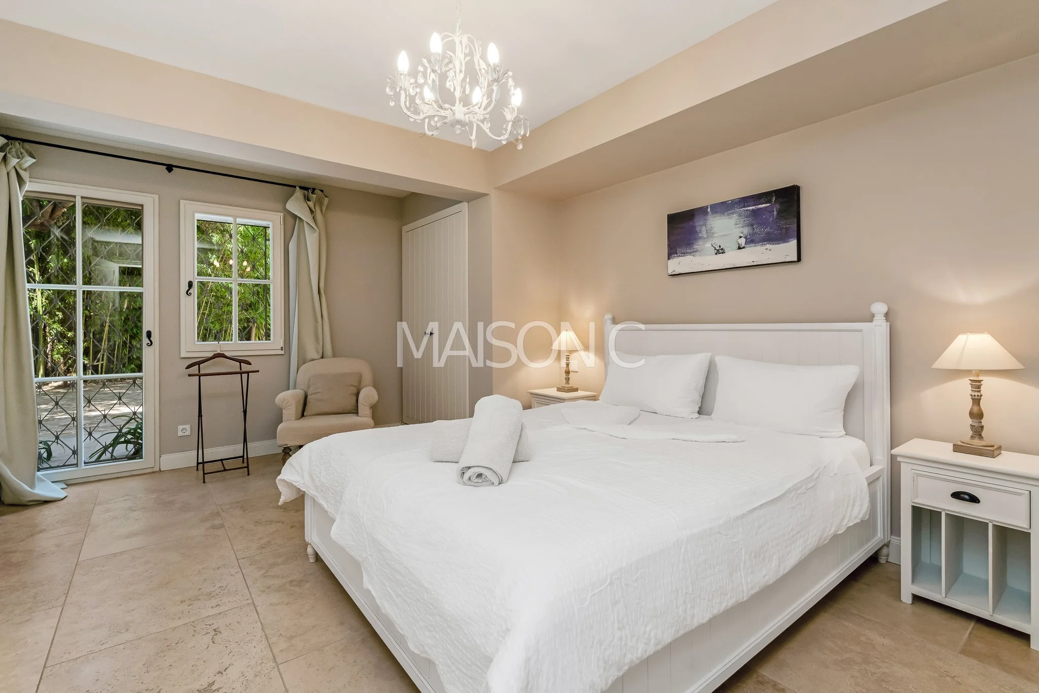 Chambre spacieuse avec lit blanc, deux lampes de chevet, tableau au-dessus du lit, fauteuil beige, fenêtres avec vue sur la nature, éclairage chaleureux.