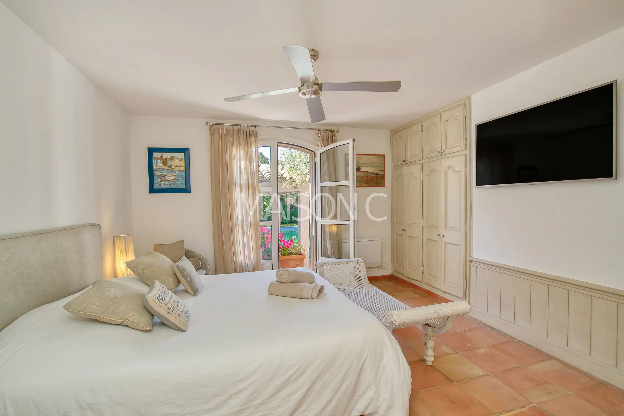 Chambre avec un lit rond, un petit fauteuil, une porte-fenêtre donnant sur un balcon avec des fleurs, un plafond avec ventilateur, un mur avec télévision et une armoire encastrée