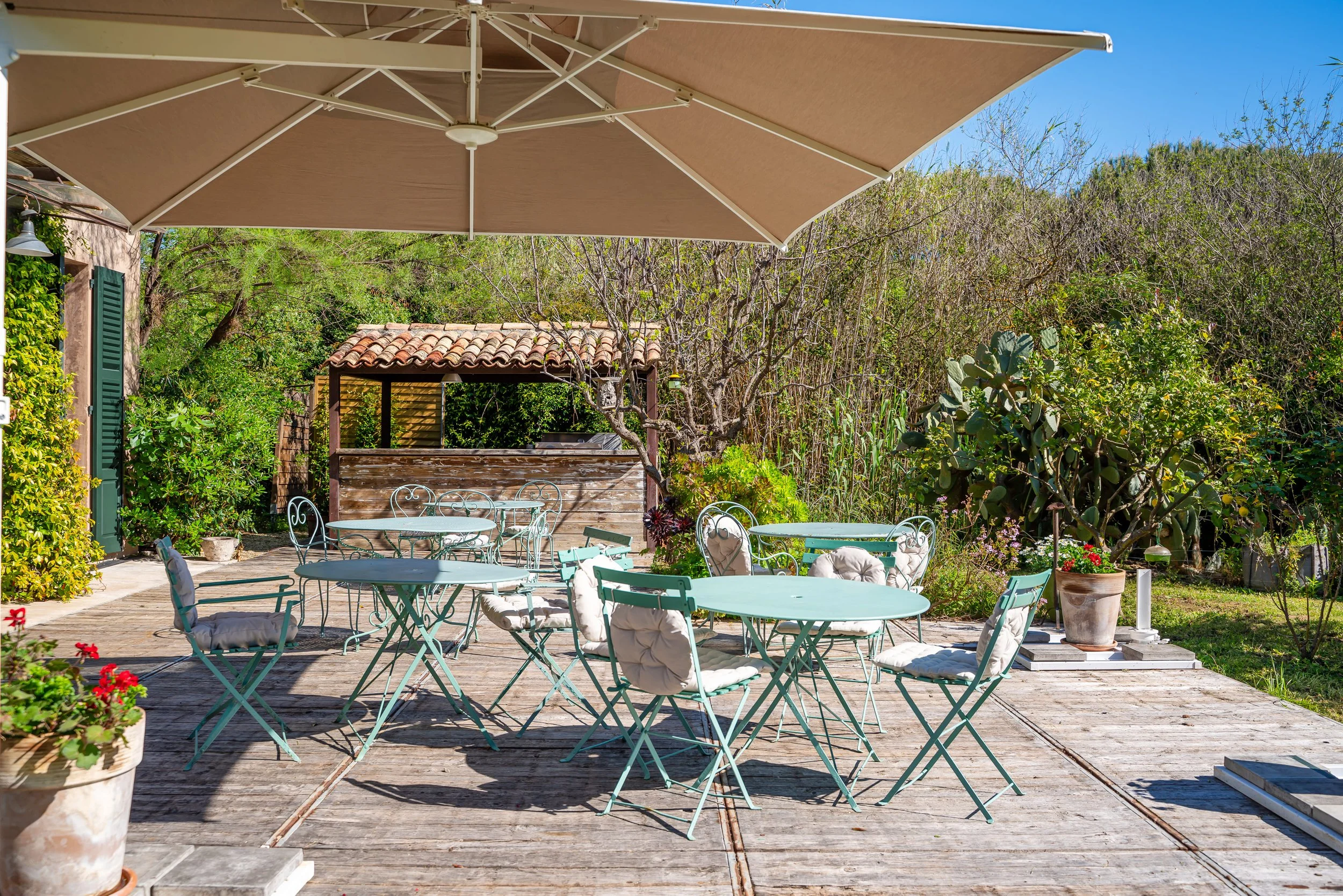 Terrasse en bois avec tables rondes en métal vert et chaises avec coussins blancs, grand parasol beige, entourée de plantes et d'arbres, espace extérieur en plein air.