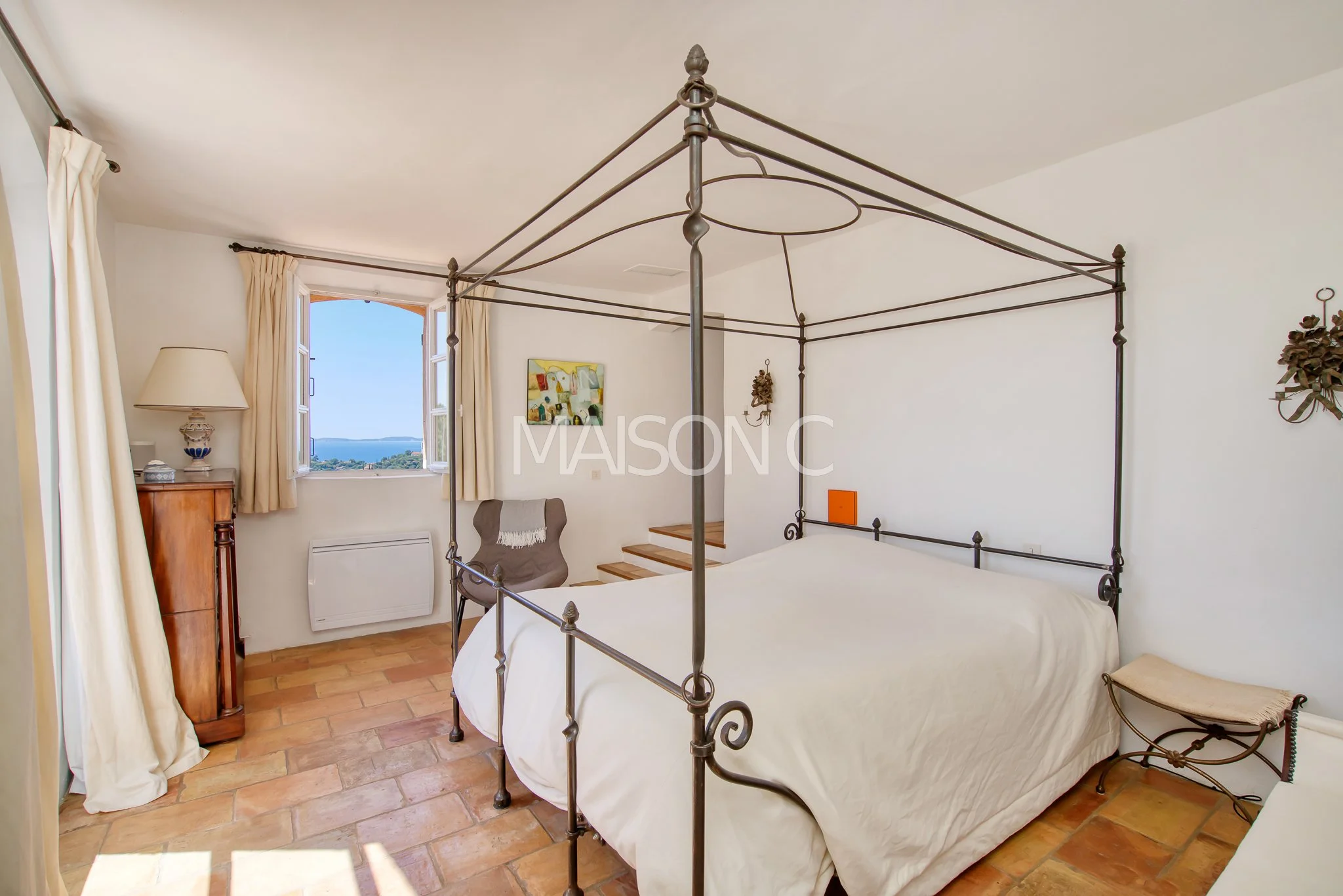 Chambre avec lit à baldaquin en métal, fenêtre avec vue sur la mer et décor vintage.
