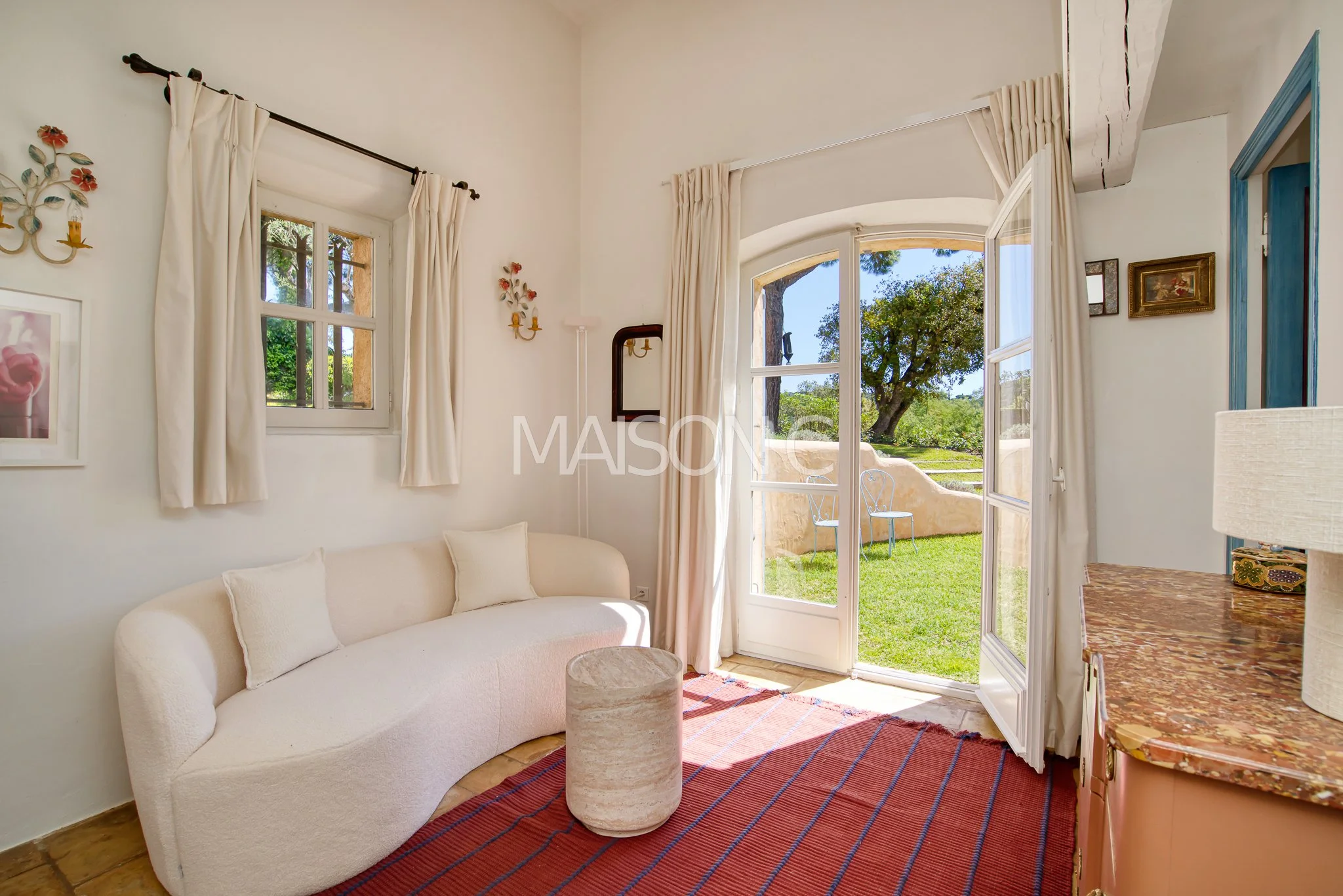 Salon lumineux avec canapé blanc, fenêtres laissant entrer la lumière naturelle, vue sur jardin avec arbres et chaises en métal, décoration bucolique.
