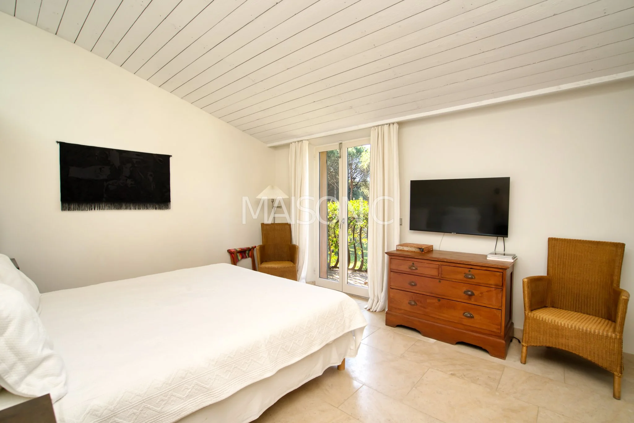 Chambre avec lit king-size blanc, télévision à écran plat sur un meuble en bois, deux fauteuils en osier, porte-fenêtre avec rideaux blancs, vue sur un balcon en nature