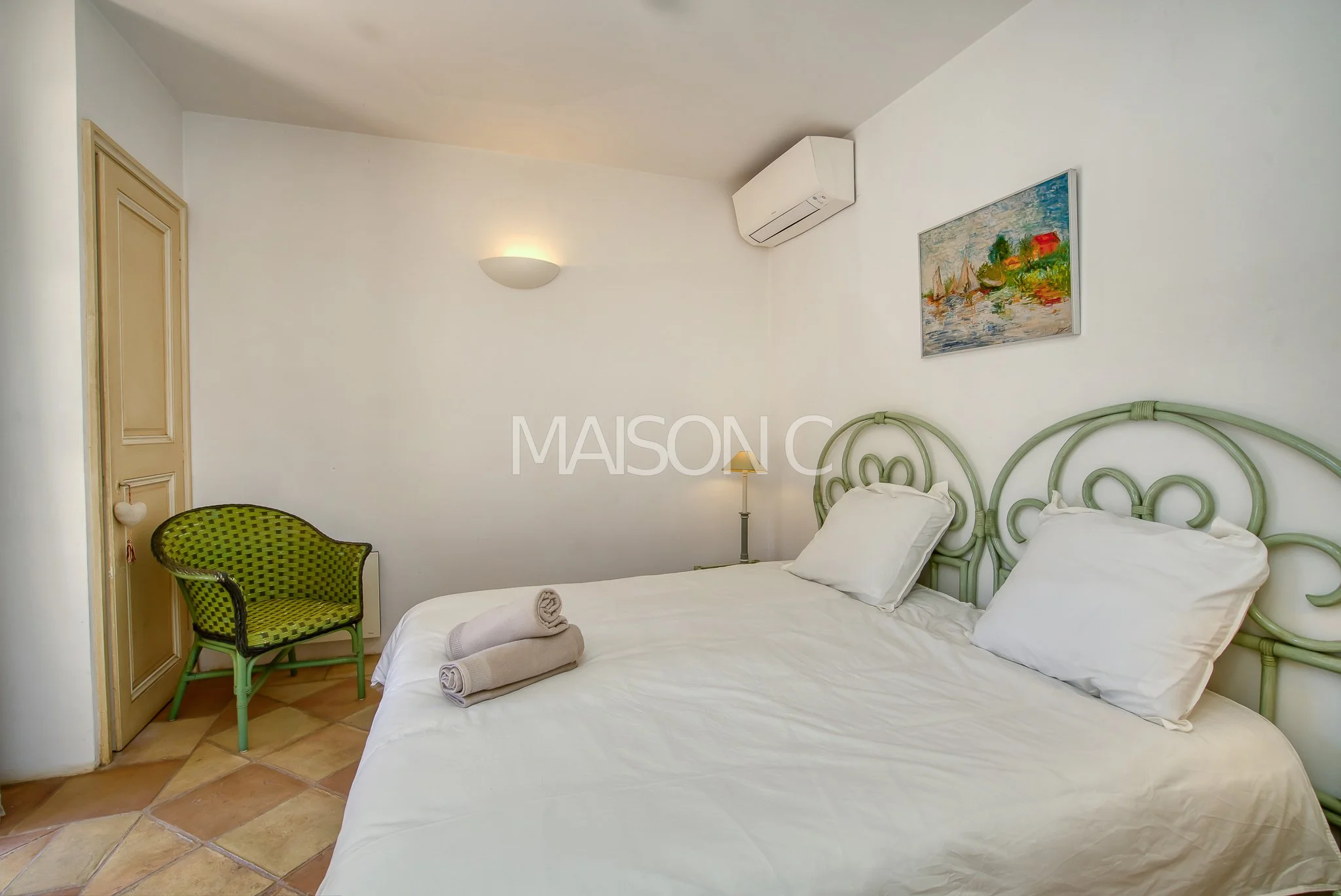 Chambre avec un lit double, deux oreillers blanc, deux serviettes roulées sur le lit, une chaise verte et un tableau coloré représentant un paysage, un climatiseur mural, et un mur avec une lumière