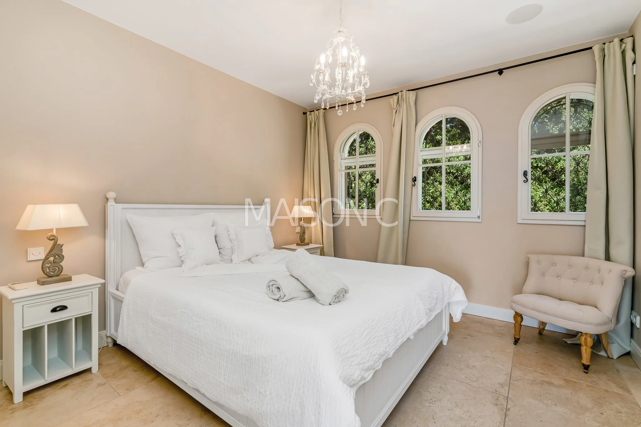 Chambre avec un lit blanc, deux tables de chevet, une lampe, trois fenêtres avec volets, un fauteuil beige, rideaux écrus, chandelier en cristaux.