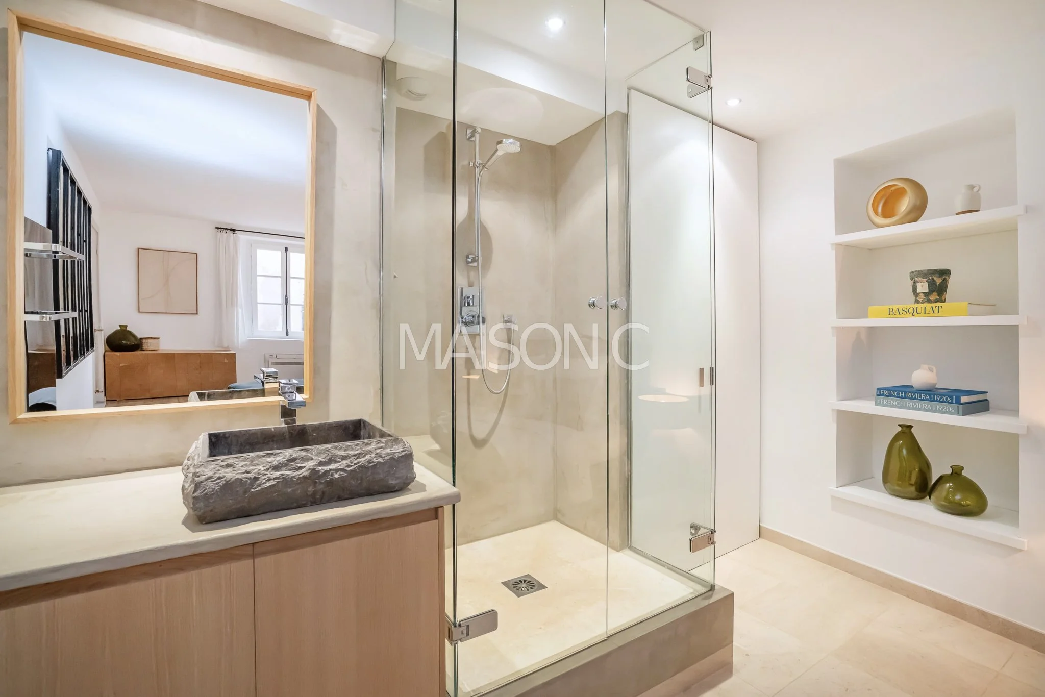 Salle de bain moderne avec douche en verre, lavabo en pierre brute, miroir et étagères décoratives avec vases et livres.