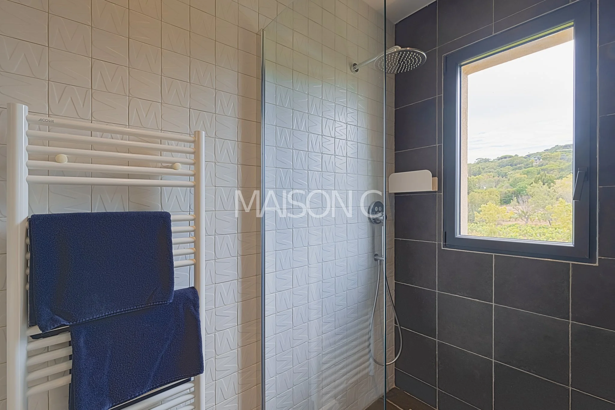 Salle de bain avec douche à l'italienne, tuiles noires et blanches, fenêtre avec vue sur la nature, serviettes bleues sur un radiateur électrique. 