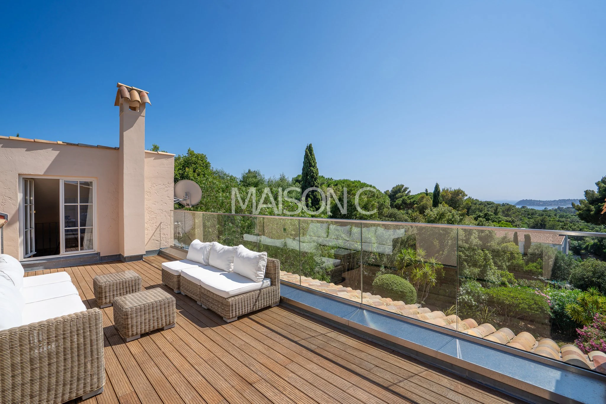 Terrasse en bois avec des meubles en rotin blanc et vue sur la nature avec des arbres et un ciel bleu clair.