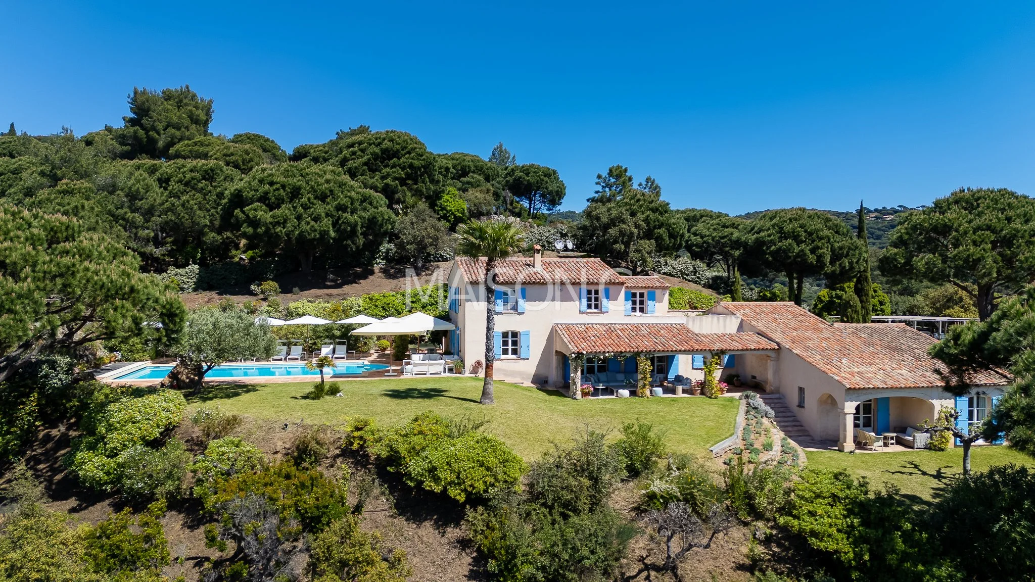 Maison de style provençal avec toits en tuiles rouges, fenêtres à volets bleus, piscine extérieure, parasols et mobilier de jardin, entourée de végétation luxuriante et de collines boisées sous un ciel bleu clair.