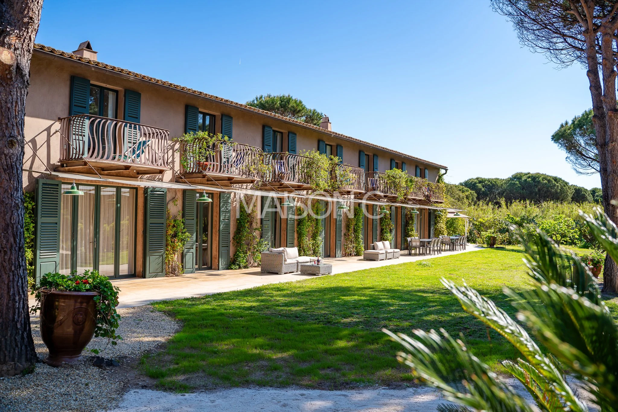 Une maison de style méditerranéen avec plusieurs balcons, entourée d'arbres et de végétation, avec un espace de détente en plein air et un ciel bleu clair.