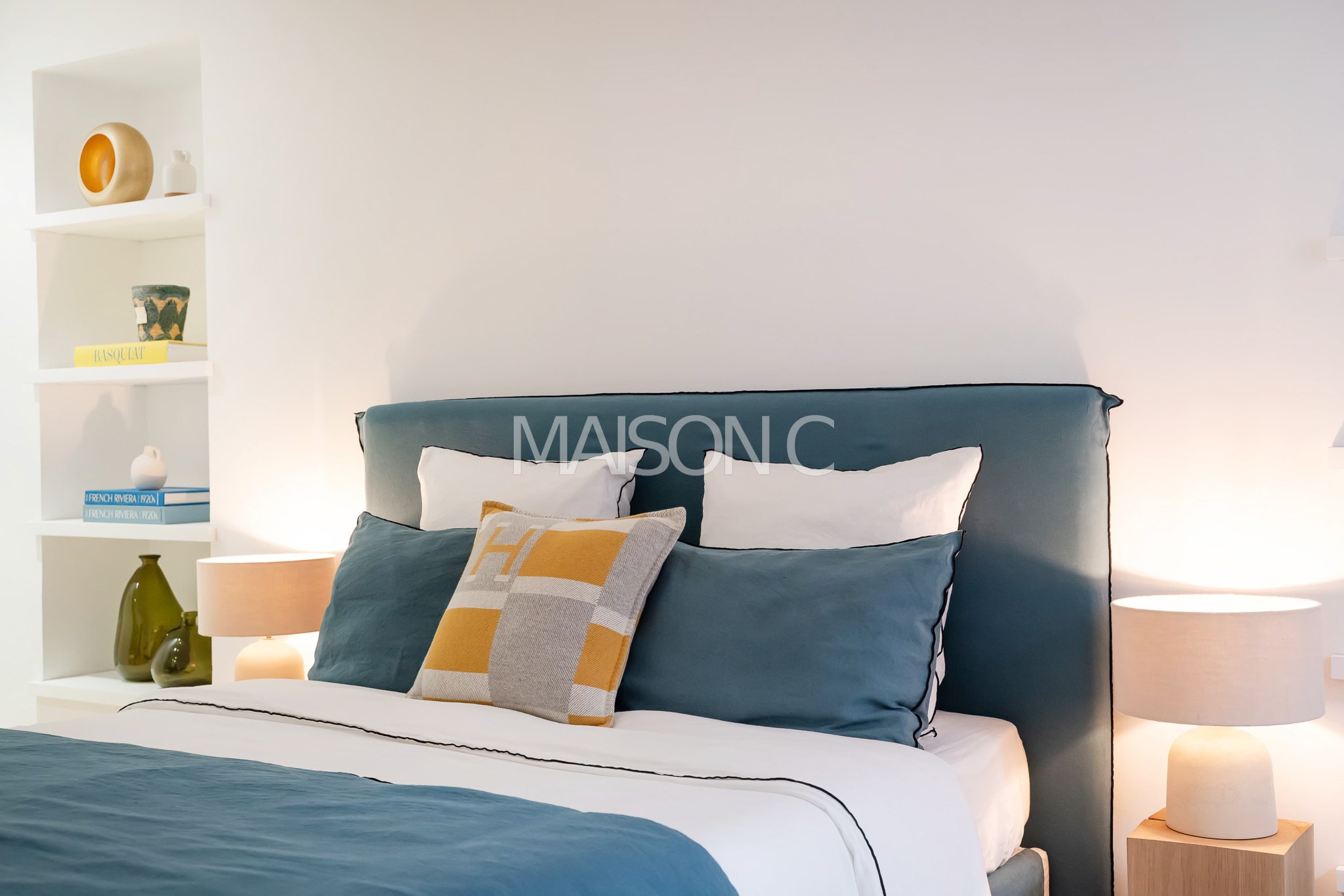 Chambre moderne avec lit à tête rembourrée, oreillers bleus et blancs, coussin décoratif jaune et gris, lampes de chevet et étagère blanche avec décorations