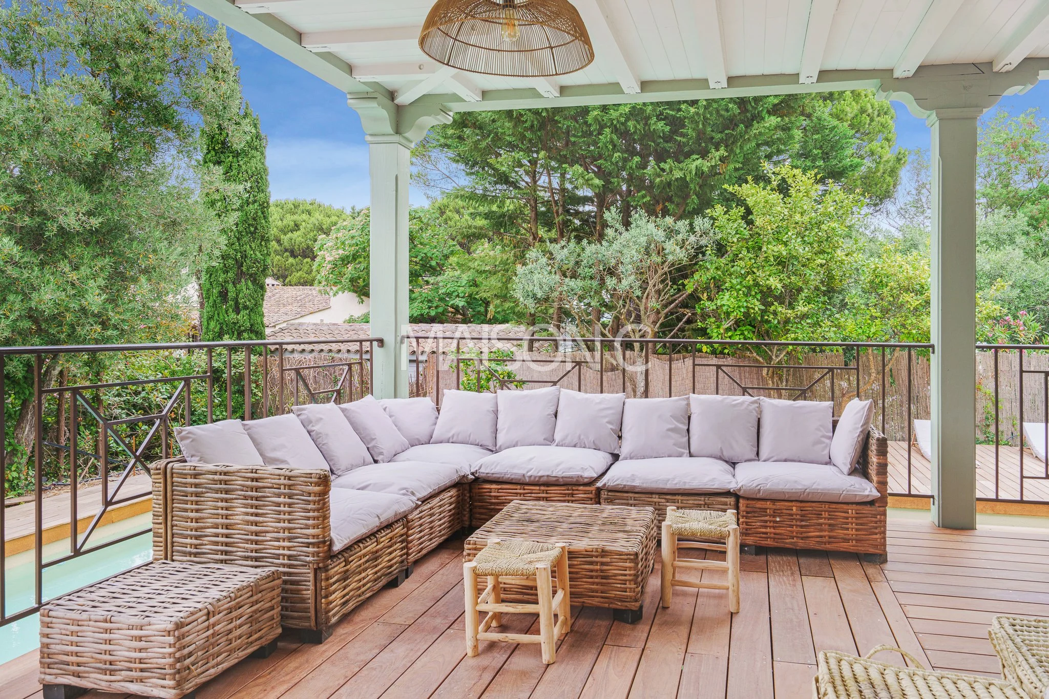 Salon de terrasse avec canapé en rotin, coussins blancs, tables et chaises en bois, arbre et ciel bleu en arrière-plan.