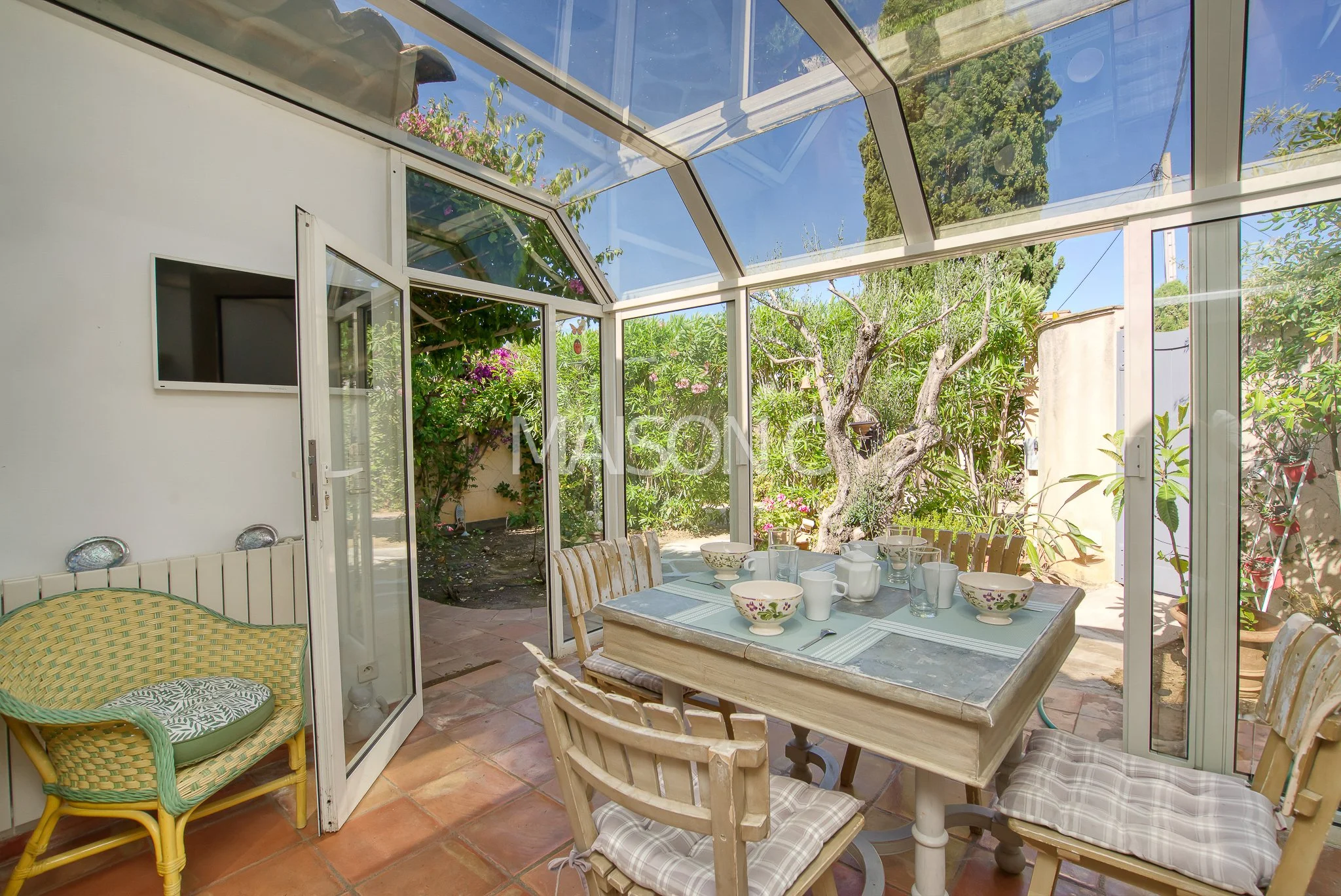 Intérieur d'une véranda avec une table à manger en bois, entourée de chaises, et des plats en porcelaine, donnant sur un jardin avec un arbre et des plantes vertes.