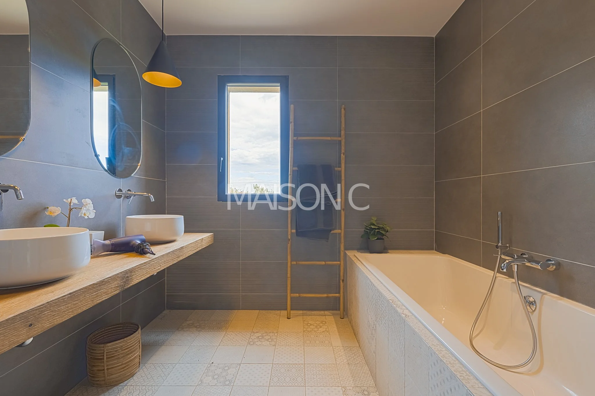 Salle de bain moderne avec double lavabo sur un plan en bois, baignoire, escalier en bois pour serviettes, fenêtre laissant entrer la lumière naturelle, murs en carreaux gris, sols à motifs, miroir rond, lampe noire et jaune, plante verte.