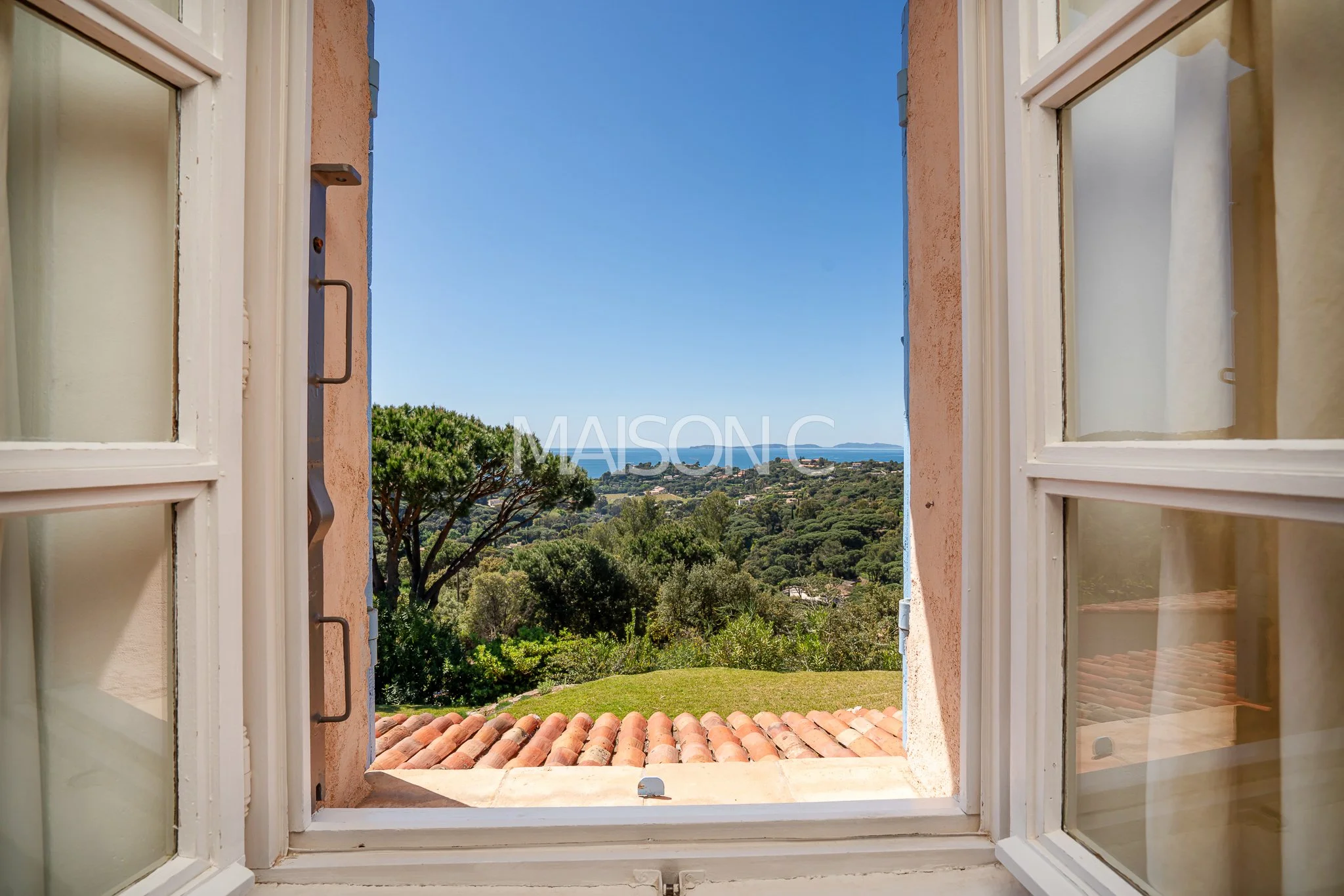 Vue depuis une fenêtre ouverte donnant sur un paysage verdoyant avec des arbres, une toiture en tuiles et la mer à l'horizon, sous un ciel bleu clair.