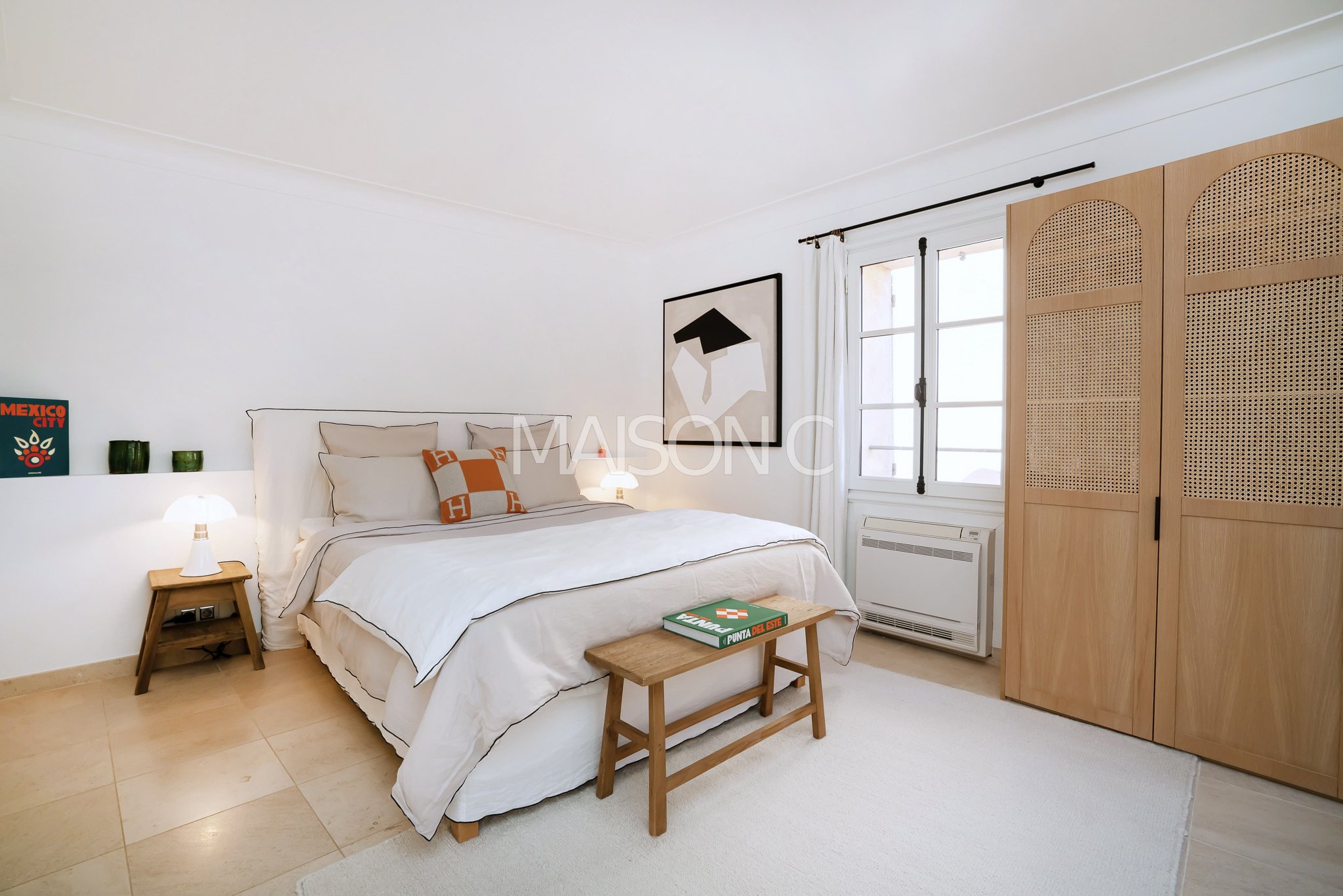 Chambre avec lit double blanc, deux lampes de chevet, tableau abstrait noir et blanc, grande armoire en bois, fenêtre avec rideaux blancs, tapis blanc, porte-manteaux avec une valise verte sur un banc en bois.