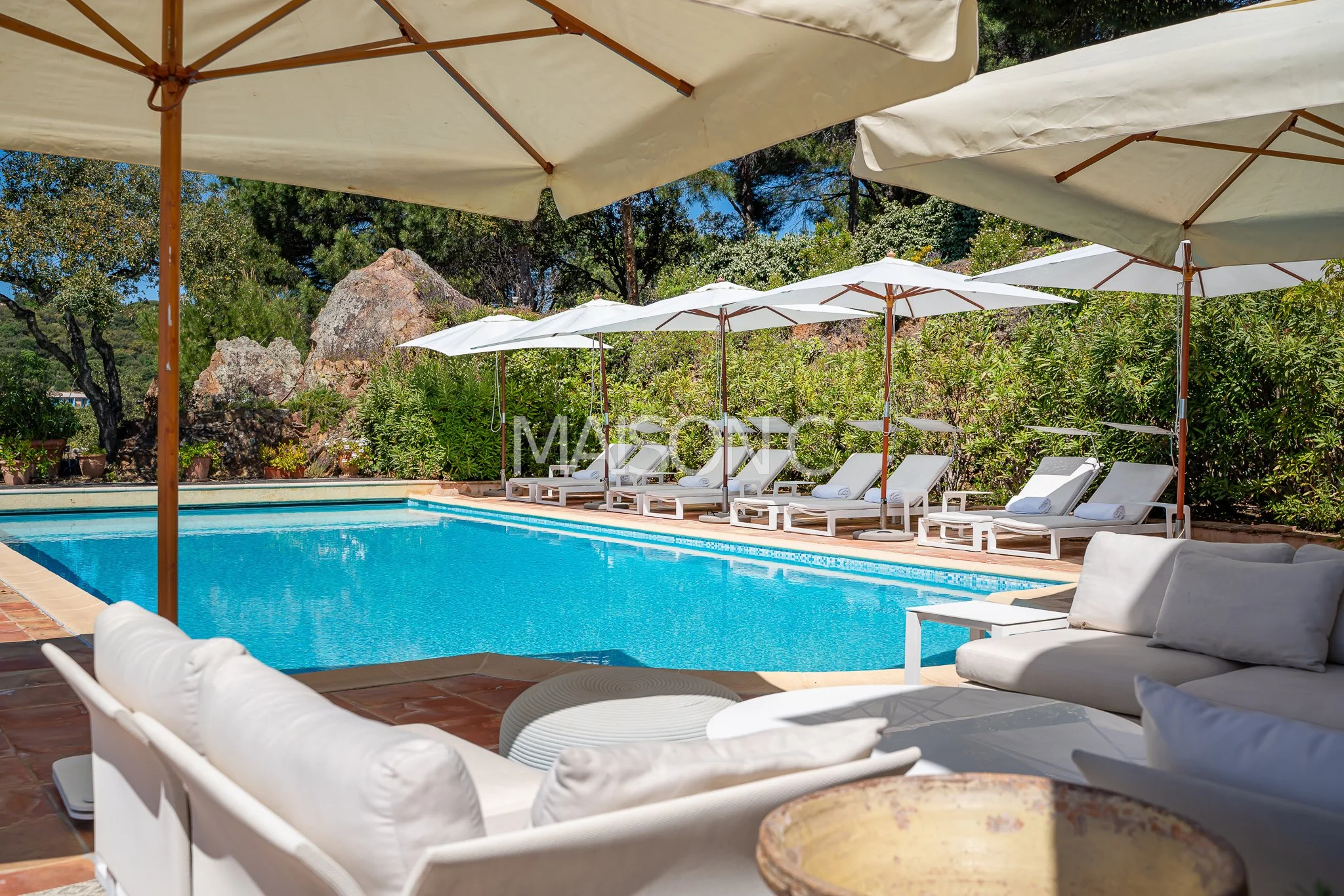 Piscine extérieure entourée de chaises longues et parasols blancs dans un jardin verdoyant.