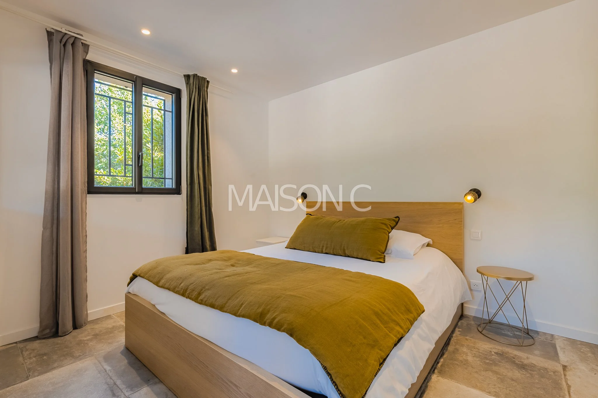 Chambre moderne avec lit en bois, couverture kaki, oreillers blancs et décor minimaliste, fenêtre avec barreaux et rideaux gris, lampe murale noire et petite table beige à côté du lit.