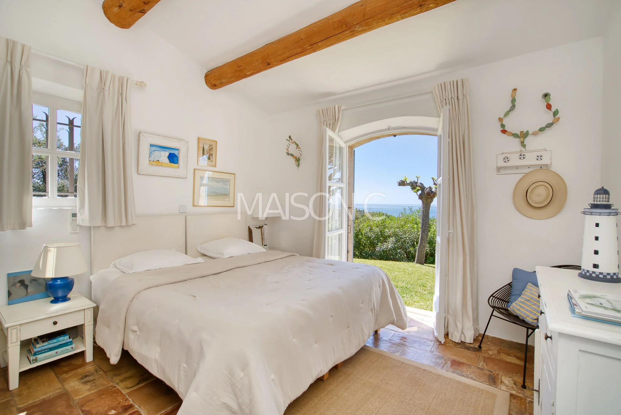 Chambre lumineuse avec lit double, décor blanc, vue sur la mer à travers une porte-fenêtre ouverte, mobilier de style coastal avec tableaux, chapeau suspendu, lampe de chevet bleue, décor marin.