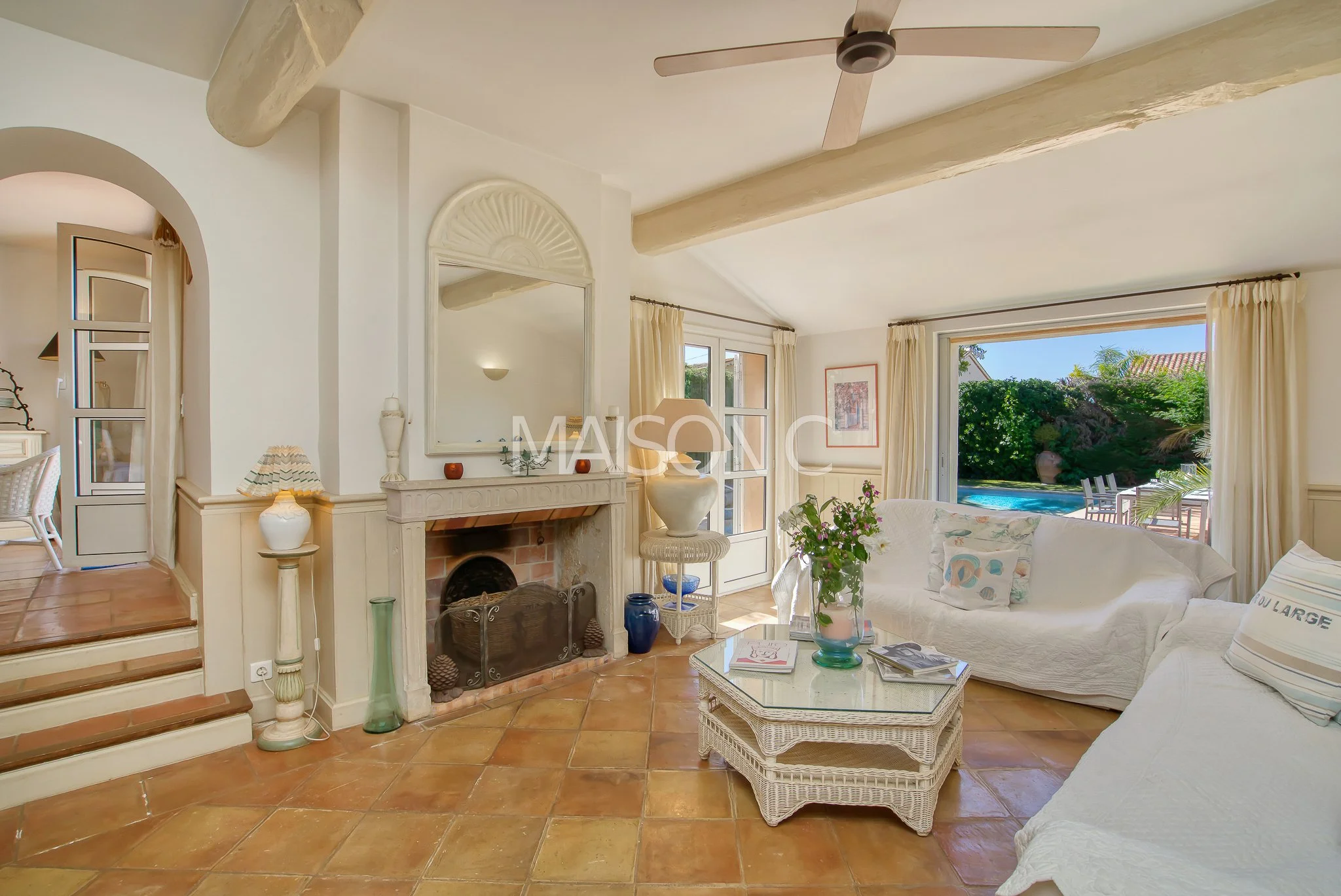 Salon lumineux avec cheminée, canapé blanc, table basse en osier, grandes fenêtres offrant une vue sur le jardin avec piscine, décoration claire et naturelle.