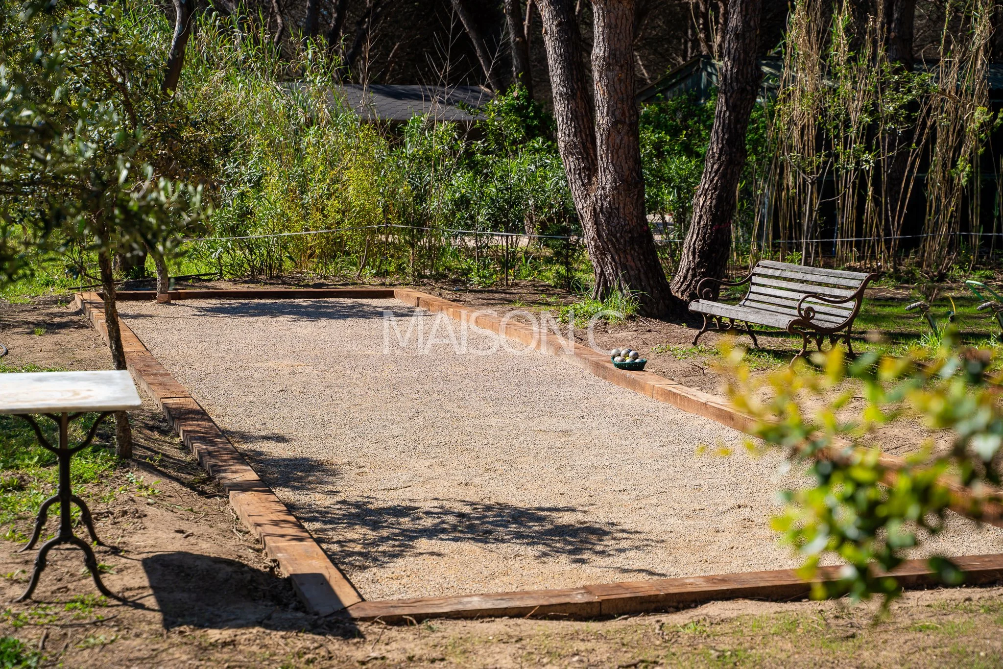 Un terrain de pétanque en extérieur, entouré d'arbres et de plantes, avec deux bancs en métal et une boîte de boules de pétanque