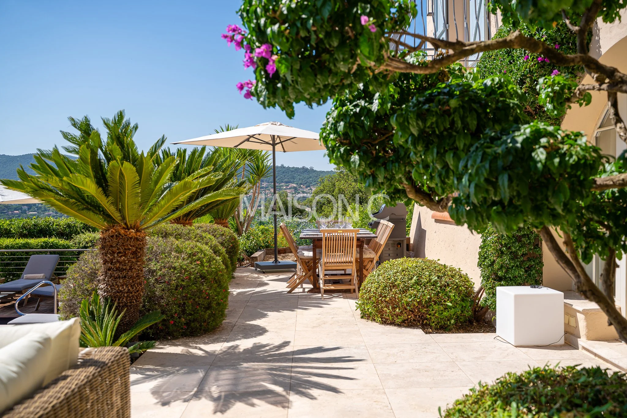 Terrasse ensoleillée avec table en bois, chaises, parasol, plantes vertes et vue sur la campagne environnante.