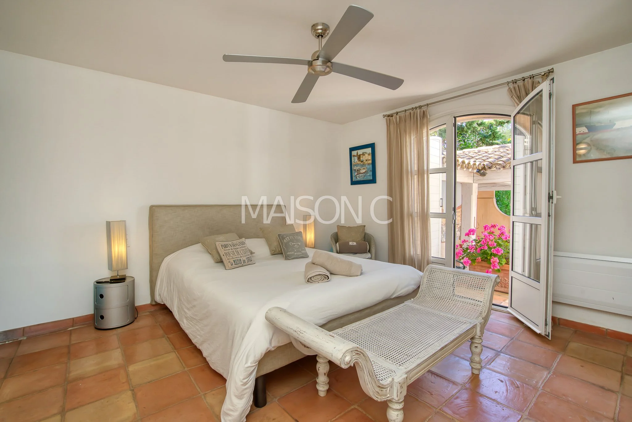 Chambre avec un lit double, un fauteuil, une table de chevet, un plafond avec ventilateur et une porte-fenêtre donnant sur un balcon avec des plantes en fleurs.